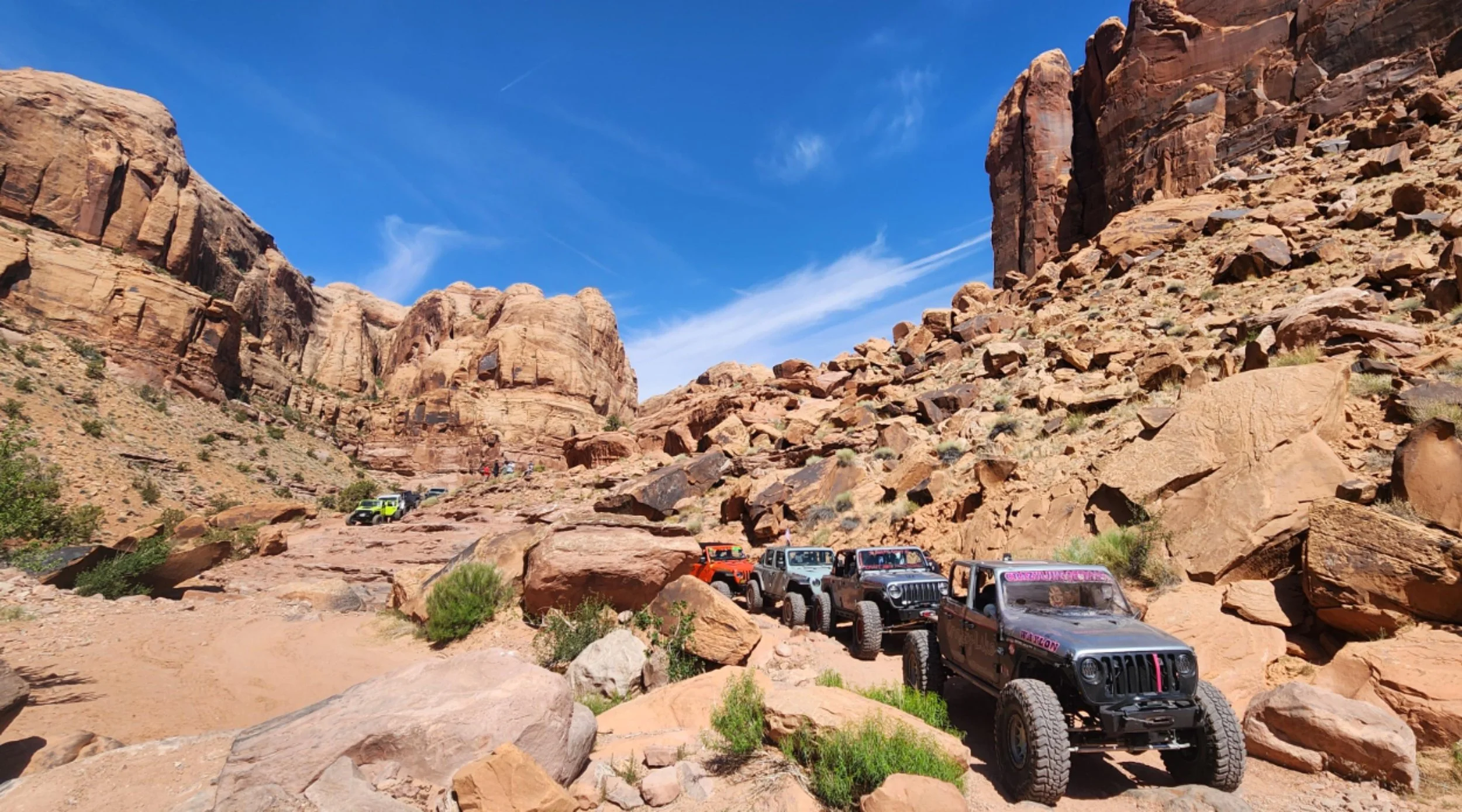 Line of off-road vehicles parked on a rocky desert trail with red rock cliffs and a blue sky in the background.