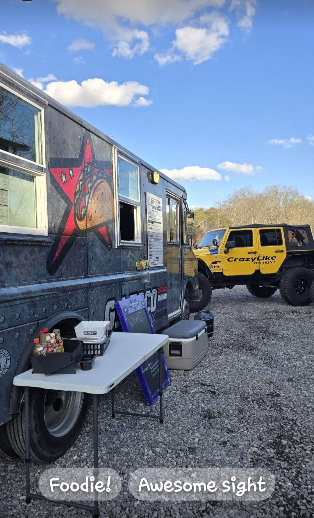Food truck with a taco star logo, a small white folding table with condiments and a black container, and two off-road vehicles, one yellow labeled 'Crazy Like Offroad'. The scene is outdoors with a gravel ground and a partly cloudy sky.