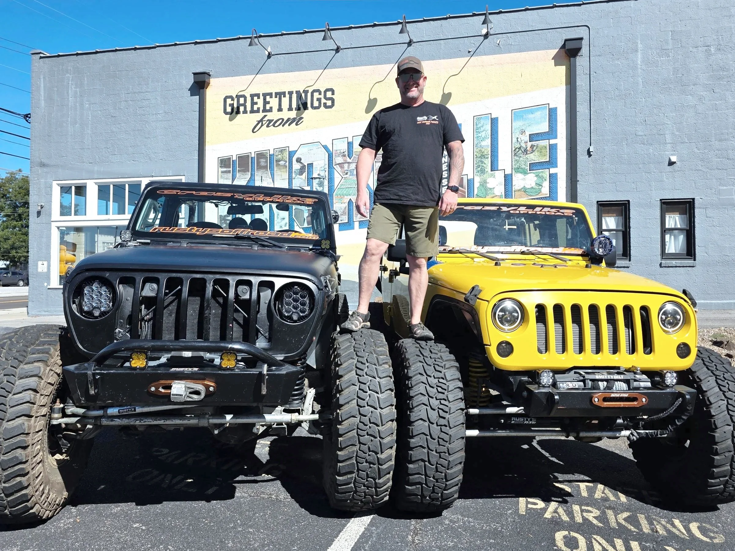 Man standing on large tires of two parked off-road vehicles in front of a mural that reads 'Greetings from Knoxville' on a building. The vehicle on the left is black and the one on the right is yellow.