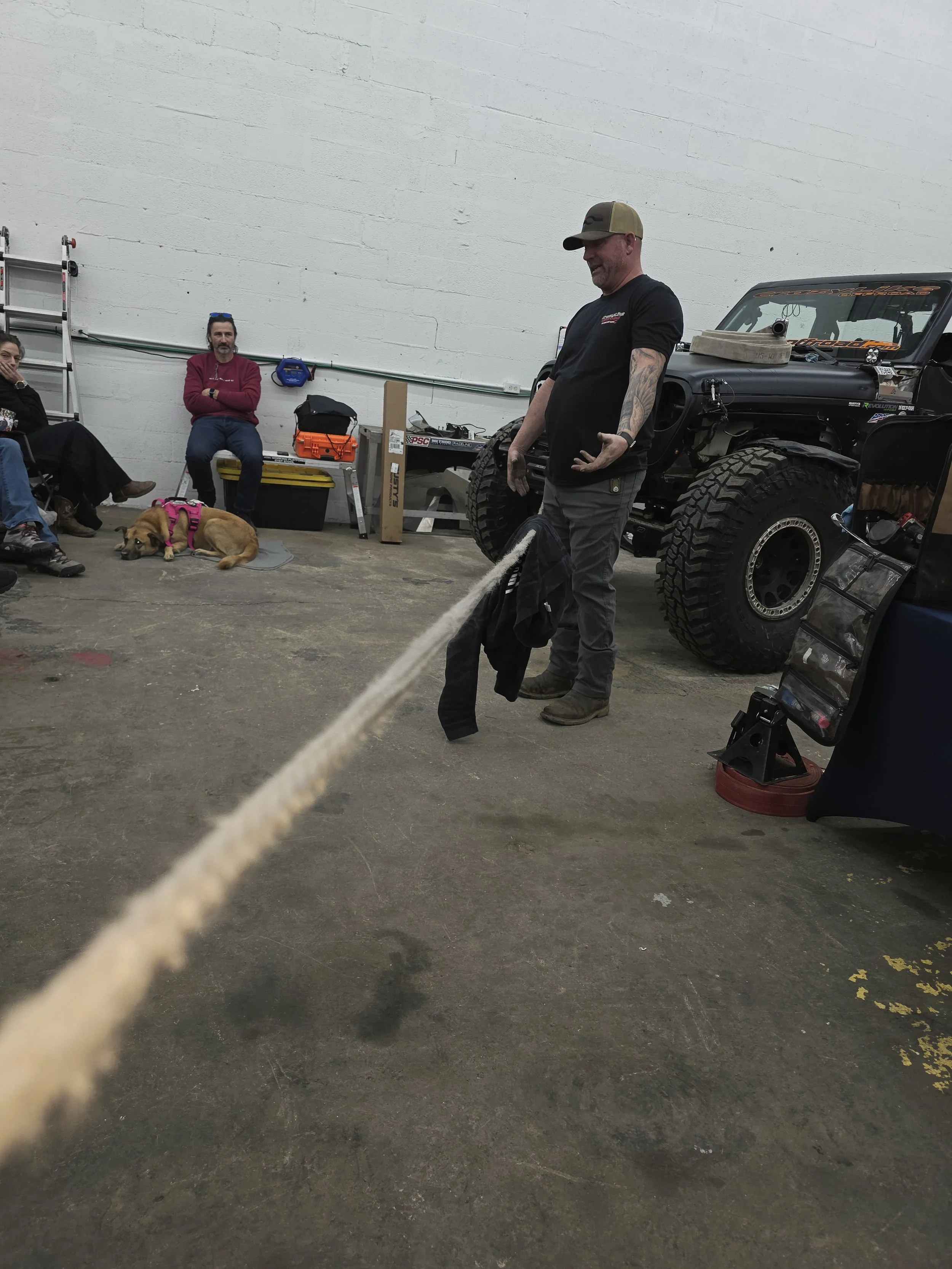 A group of people sitting on a bench and on the floor in a garage or workshop, with a dog lying down, and a man standing in front of a black off-road vehicle showing a rope.