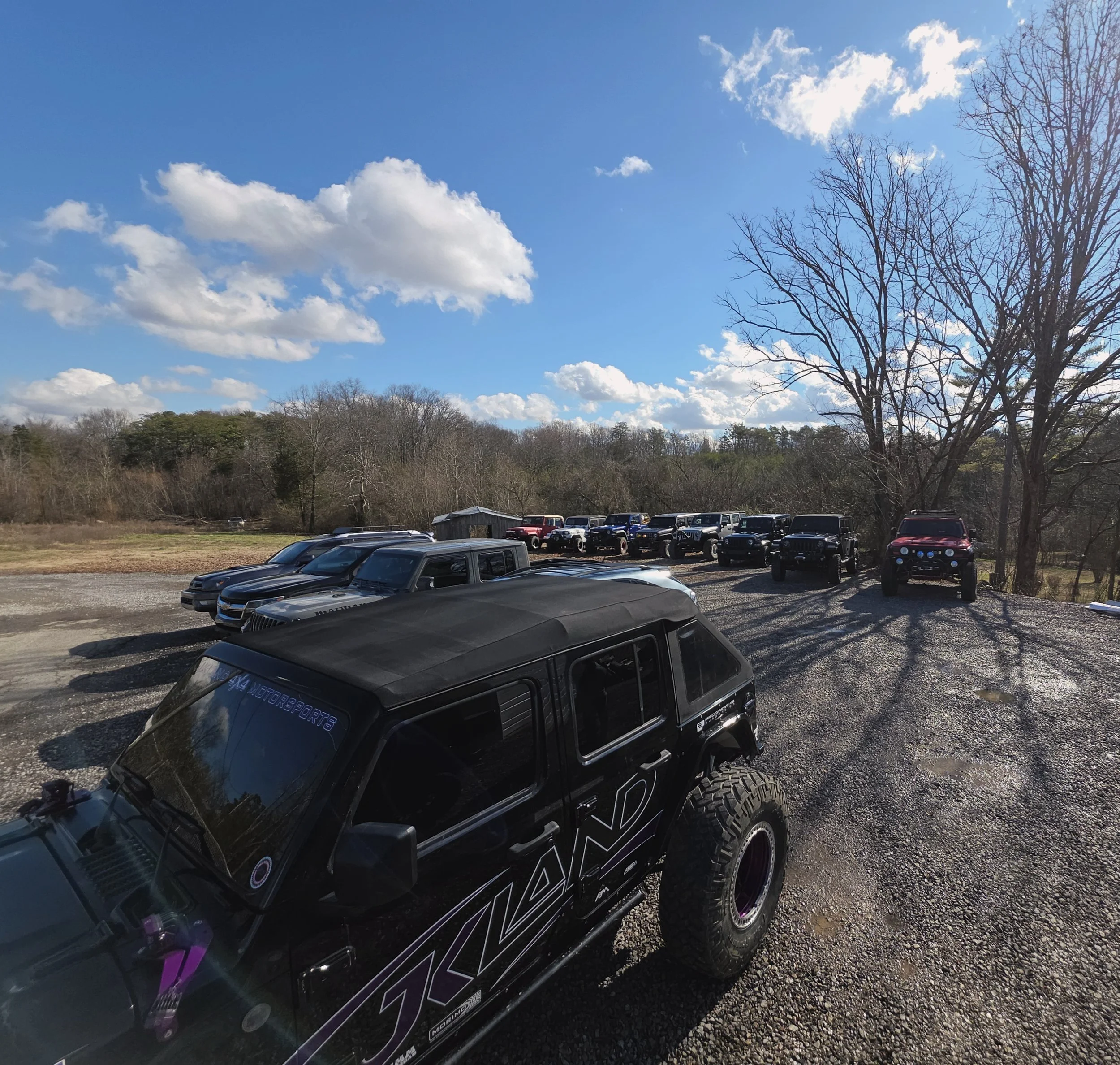 A gravel parking lot filled with various dark-colored trucks and SUVs, some with off-road modifications, on a partly cloudy day with blue sky and scattered clouds, surrounded by bare trees and a hilly wooded background.