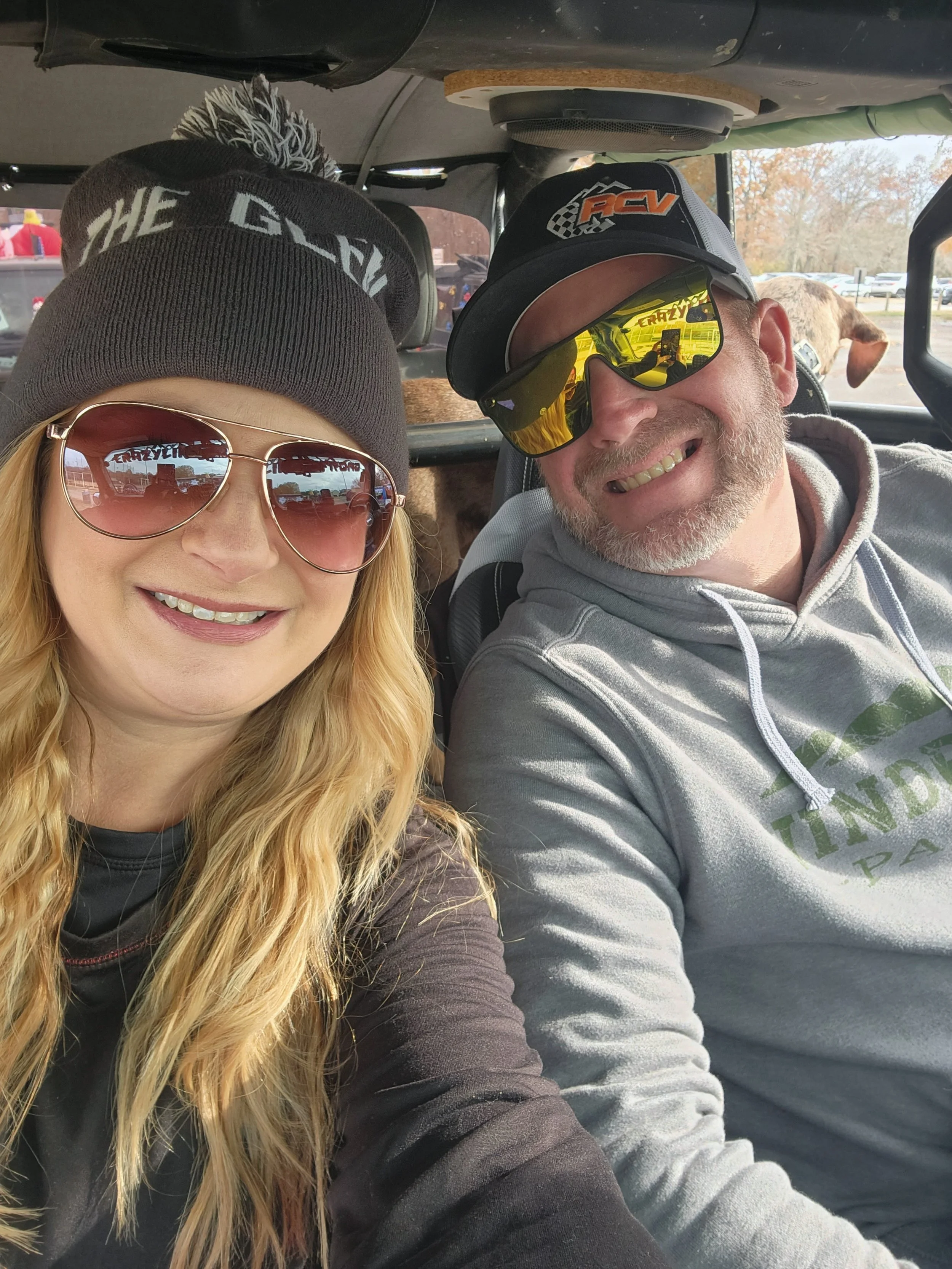 A woman and a man sitting in a vehicle, smiling for a selfie. The woman is wearing sunglasses and a beanie, while the man is wearing a baseball cap and reflective sunglasses. There are horses visible in the background outside of the vehicle.