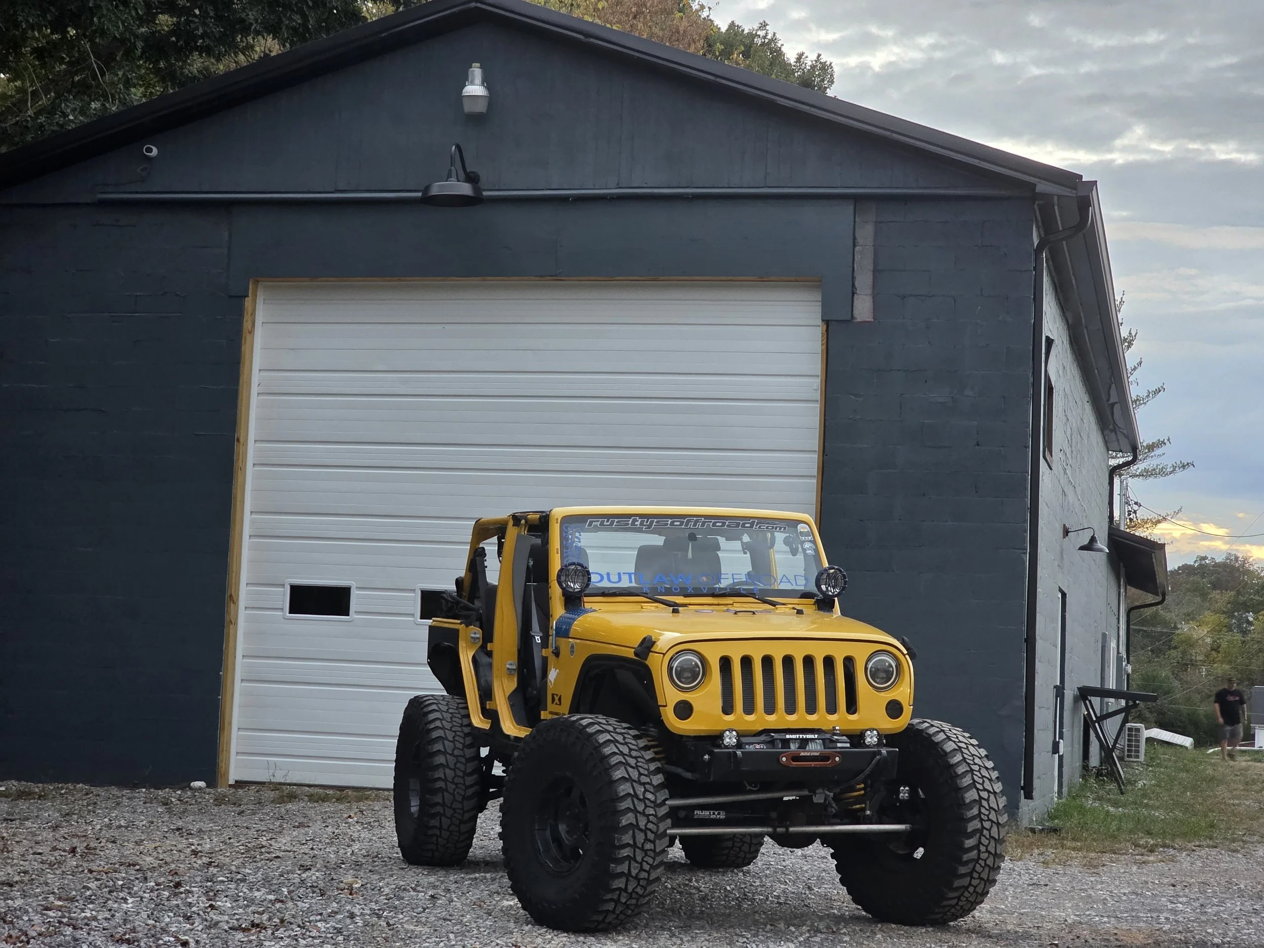 Yellow off-road Jeep with large tires parked in front of a dark gray warehouse with a large white garage door and a small side door. A person is walking in the background to the right.