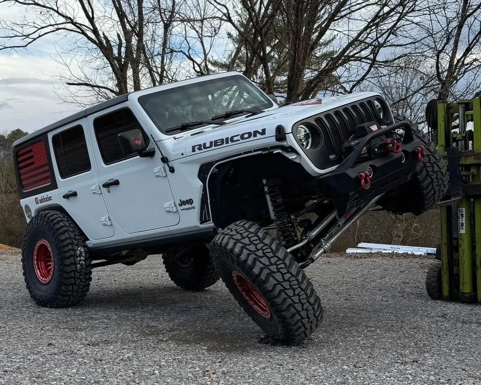 A white Jeep Rubicon with red accents being lifted on a forklift, with its front left wheel elevated. The vehicle has off-road tires, black grille, and decals on the side and rear window, with a background of trees and a partly cloudy sky.