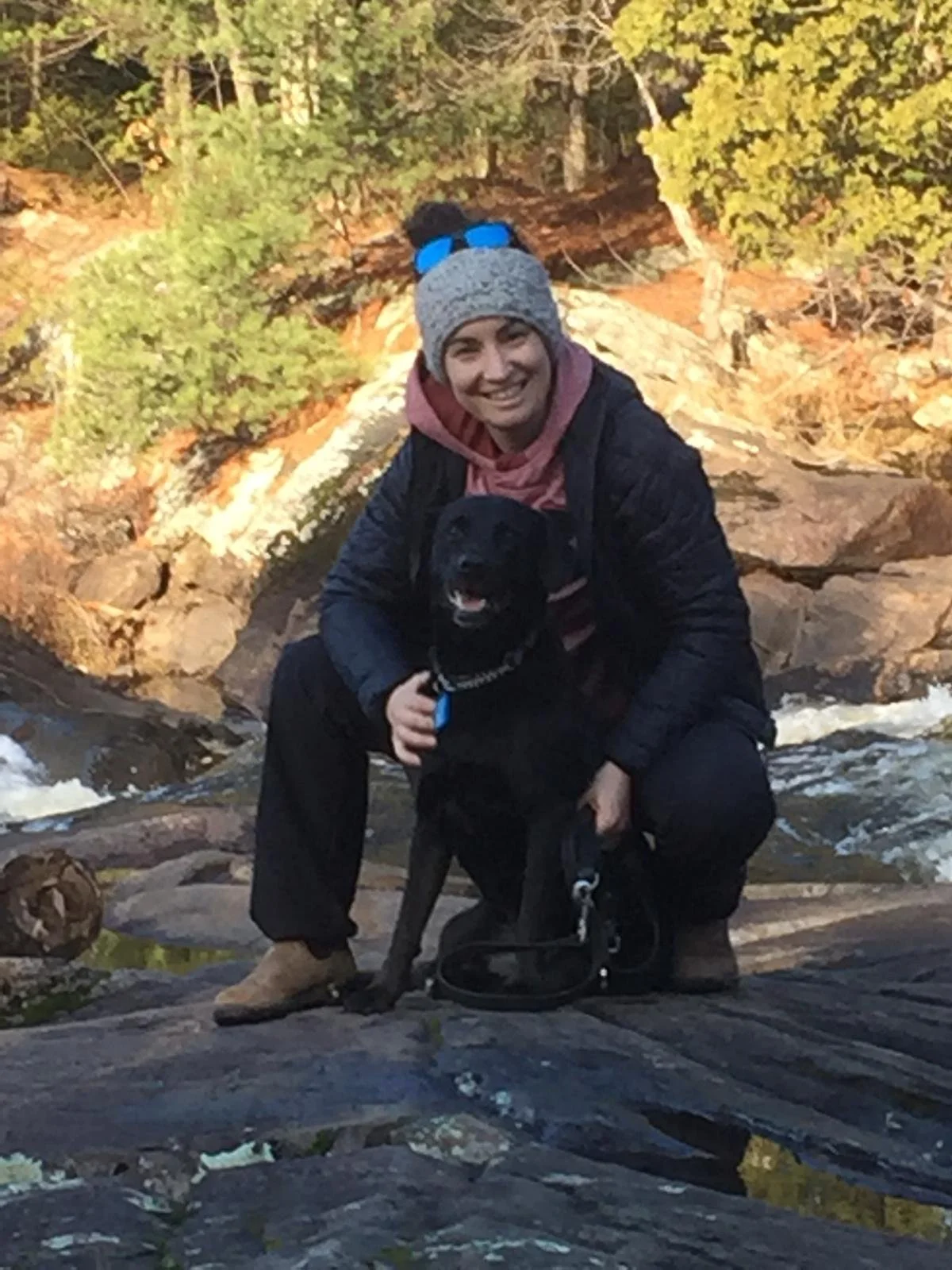 A woman with a gray knit hat, pink hoodie, black jacket, and tan boots outdoors on rocks, smiling while holding a black dog with a blue collar near a flowing stream and surrounded by trees with autumn foliage.