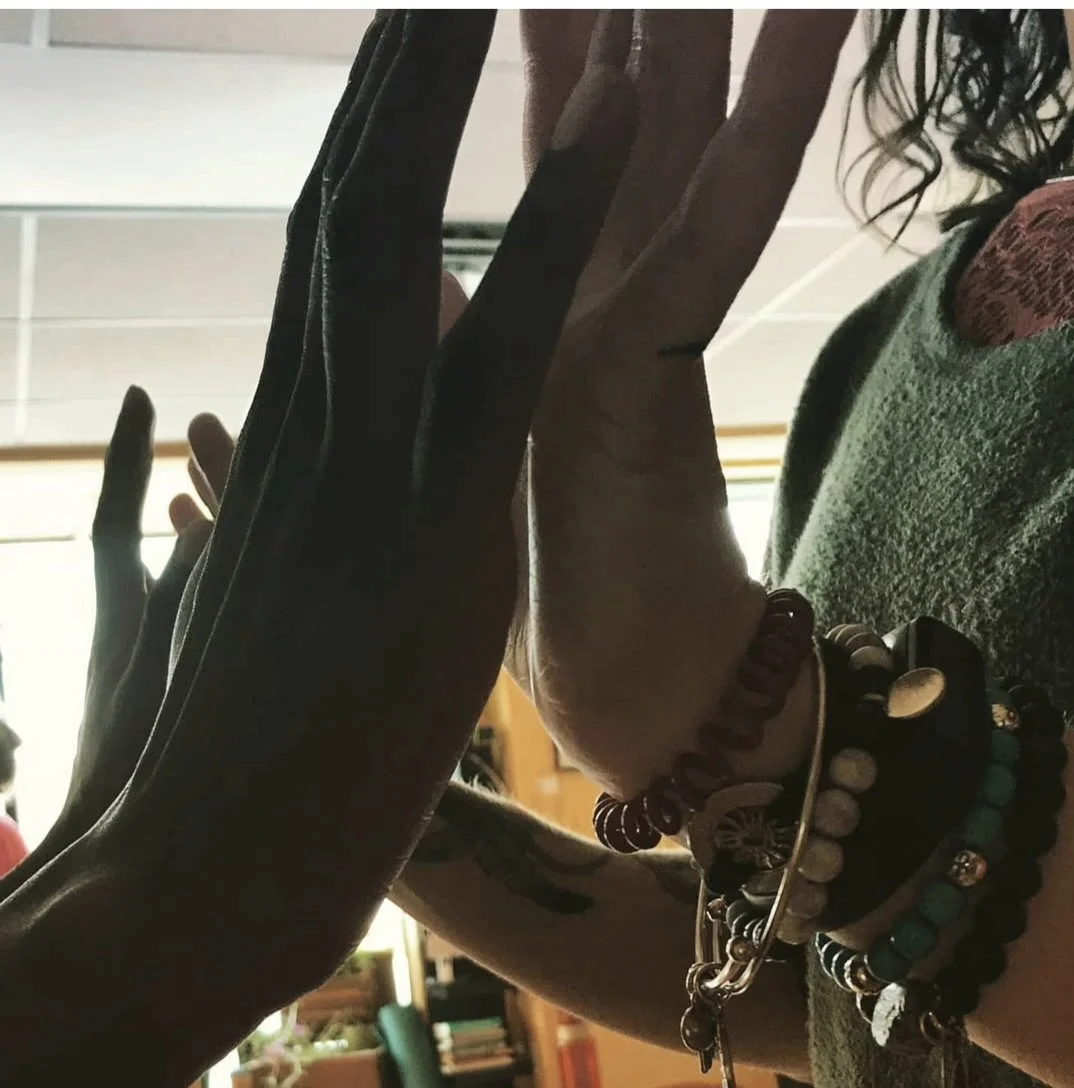 Two people holding their hands energetically just slightly apart, palms facing, with several bracelets and tattoos visible on their wrists.