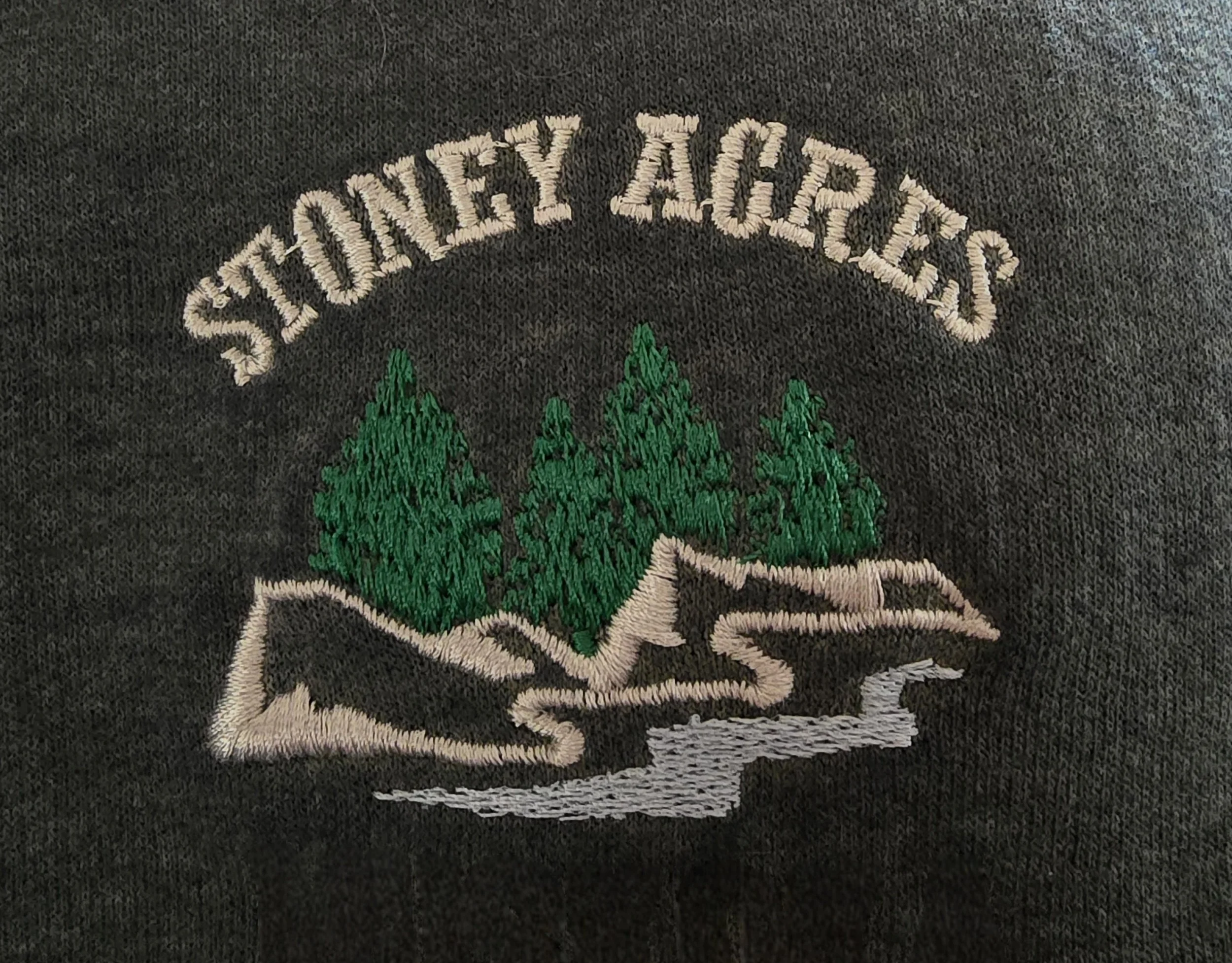 Embroidered logo for 'Stoney Acres' featuring a stylized river flowing past rocks and three green pine trees on black fabric.