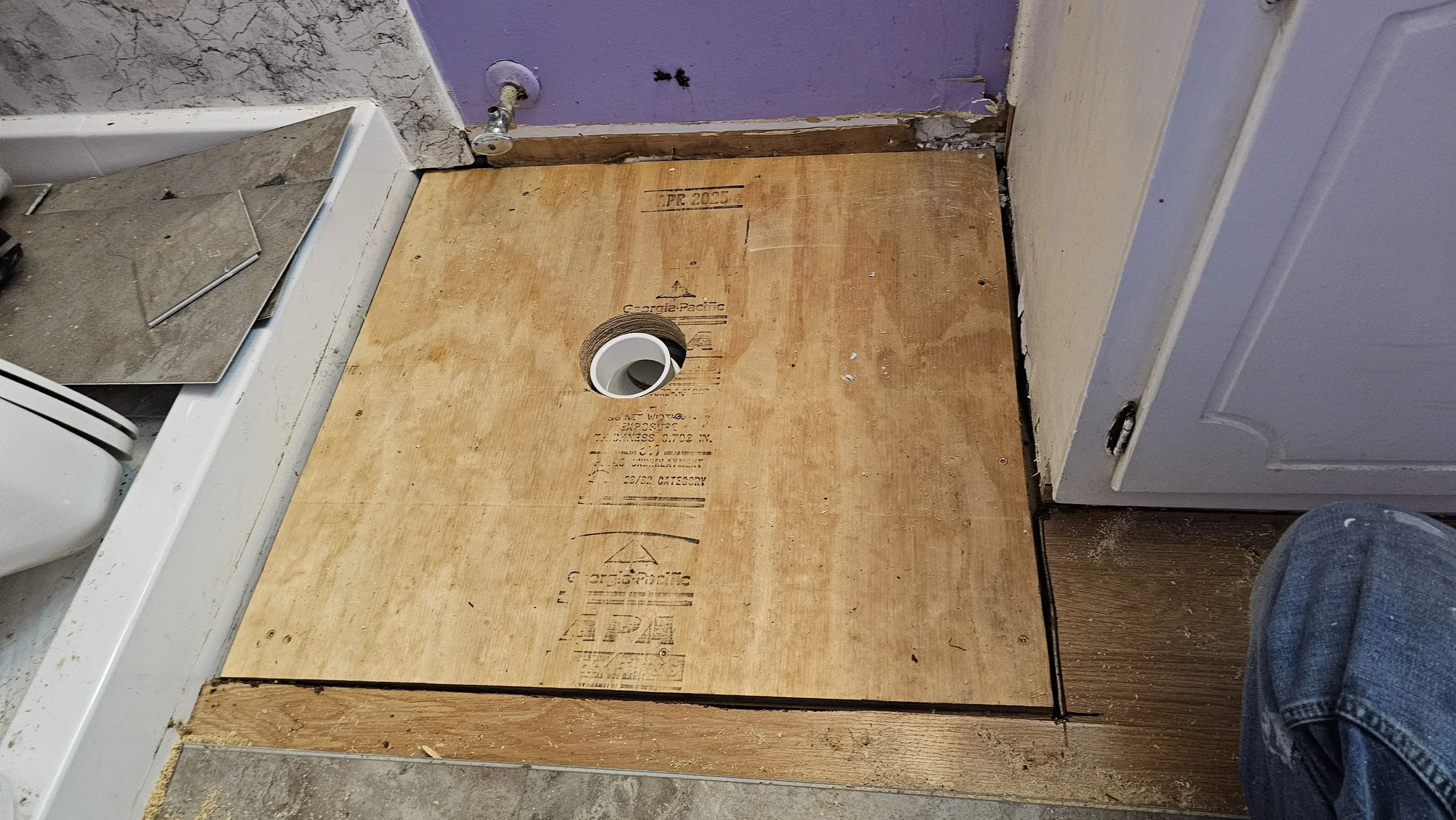 Bathroom floor with plywood subfloor installed. A circular hole is cut in the plywood around a white PVC pipe. A mirror and some tiles are visible on the left, and a washing machine or dryer is on the right. A purple wall with exposed pipes and trim 