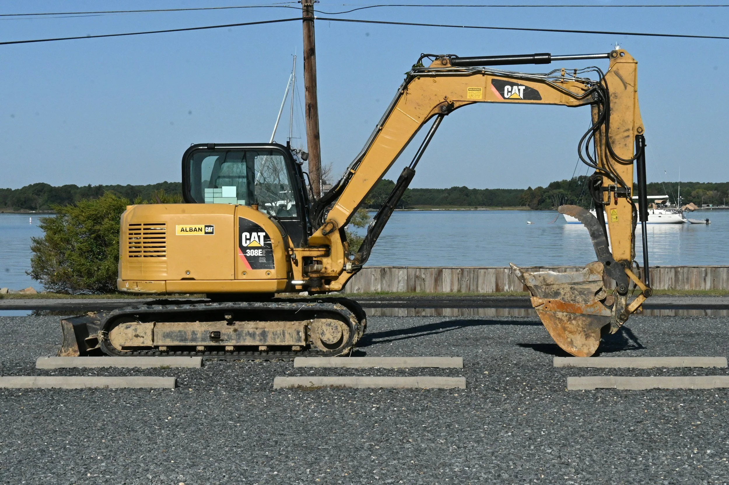 Yellow CAT excavator parked on gravel, with water and sailboats in the background.