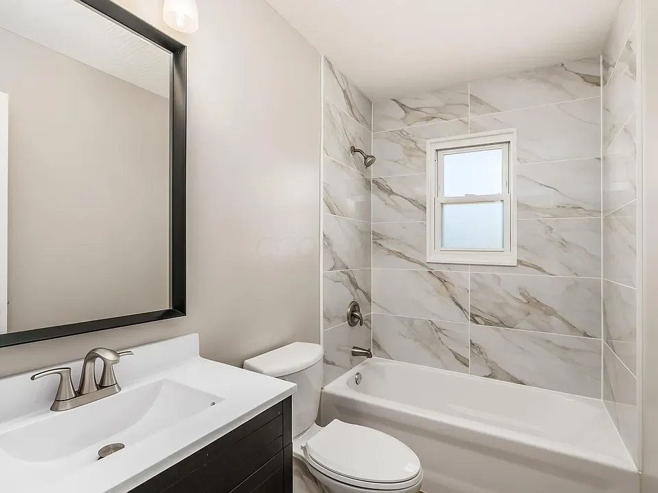 Modern bathroom with a vanity, mirror, toilet, bathtub, and a small frosted window, featuring marble wall tiles.