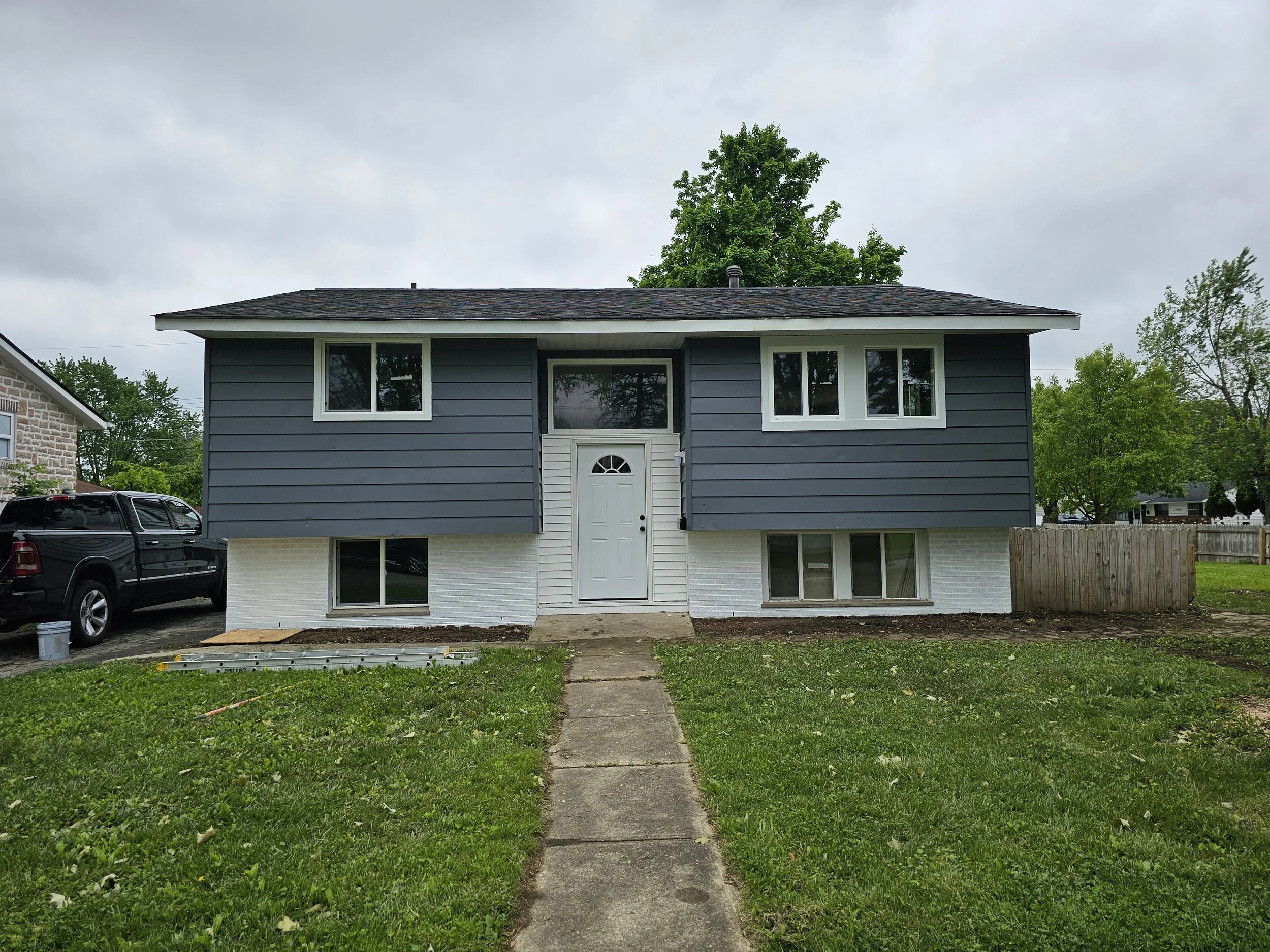 Front view of a two-story house with dark blue siding and a white basement, with a concrete walkway leading to the front door and a black truck parked on the driveway.