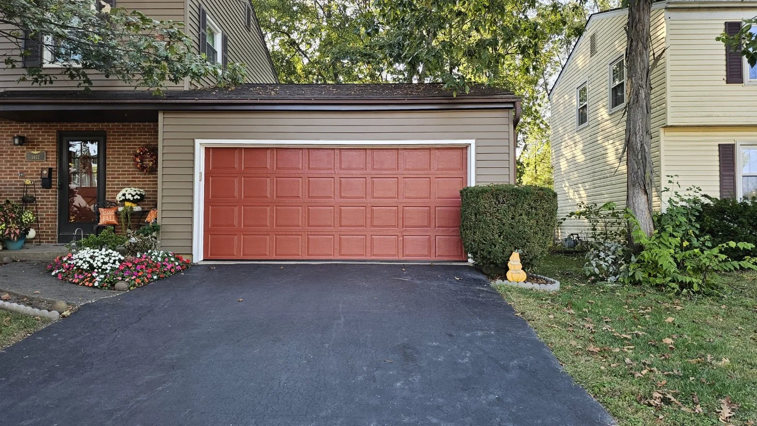 Front view of a house with a red garage door, surrounded by flowers, plants, and decorative items, with neighboring houses on either side and trees overhead.