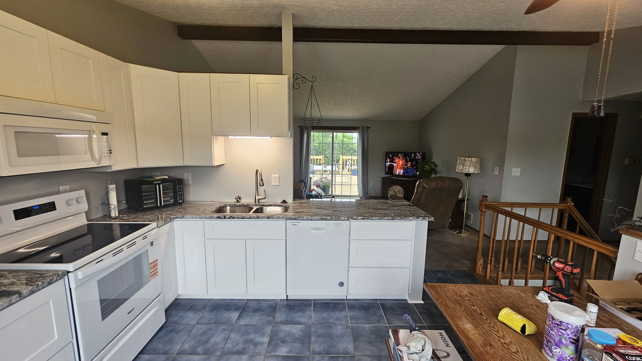 Kitchen with white cabinets, granite countertops, stove, microwave, toaster, sink, and a view into the living room with a TV and a sliding glass door leading outside. A wooden table with a drill, paper towel, and other items is in the foreground.