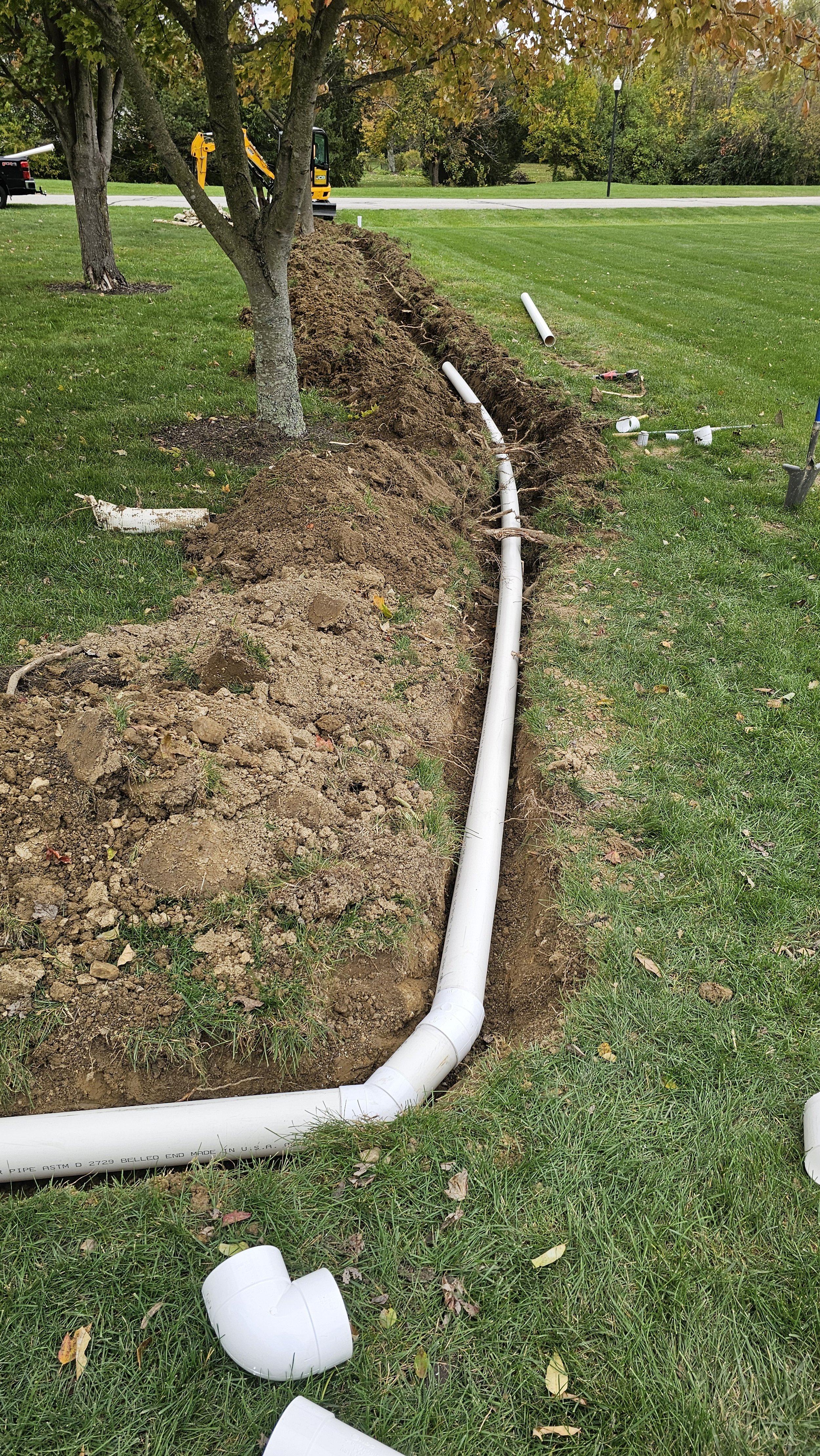 PVC pipes are being installed underground in a grassy area as part of a landscaping or construction project. An excavator and tools are visible in the background.