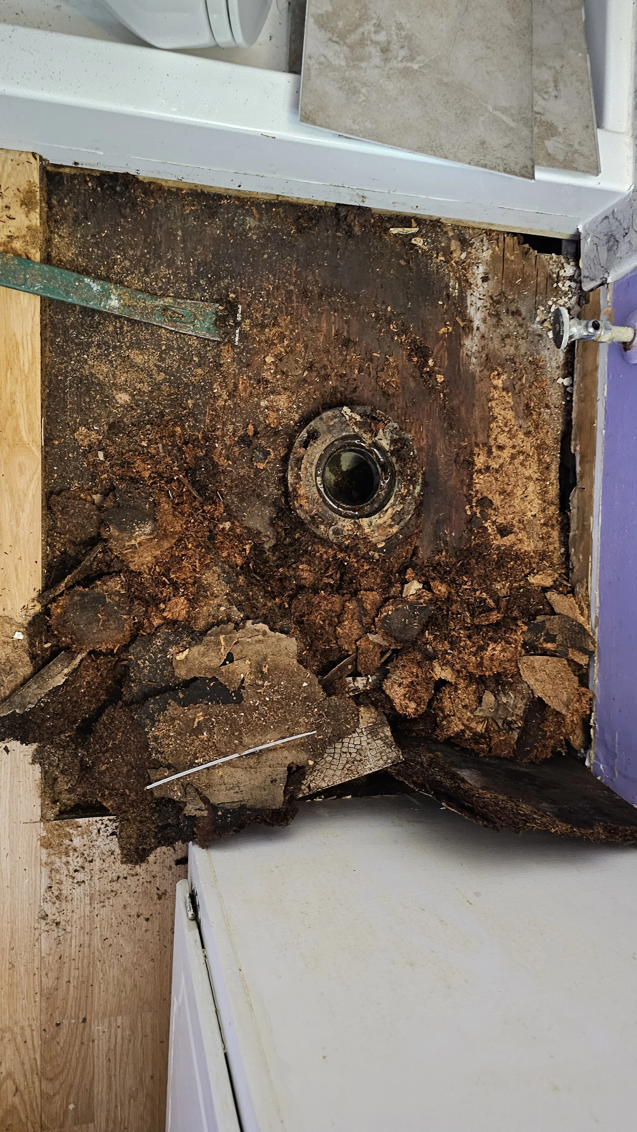 Kitchen floor with removed dishwasher, exposing a rusty drain pipe and debris.
