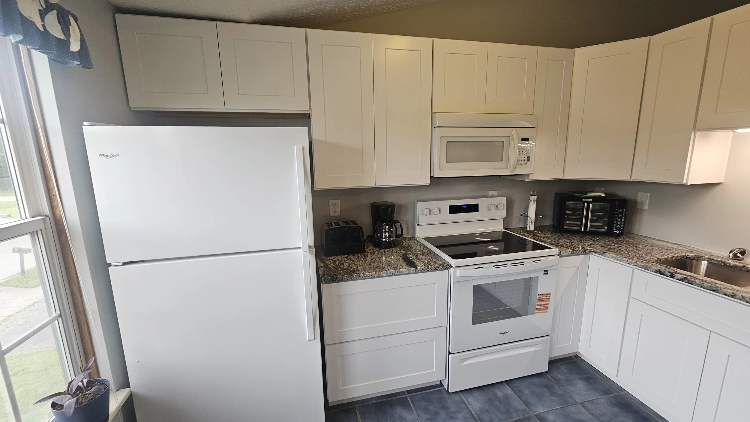 Kitchen with white appliances including a refrigerator, microwave, oven, and toaster, beige upper and lower cabinets, granite countertops, a double sink, and a window with a blue curtain.