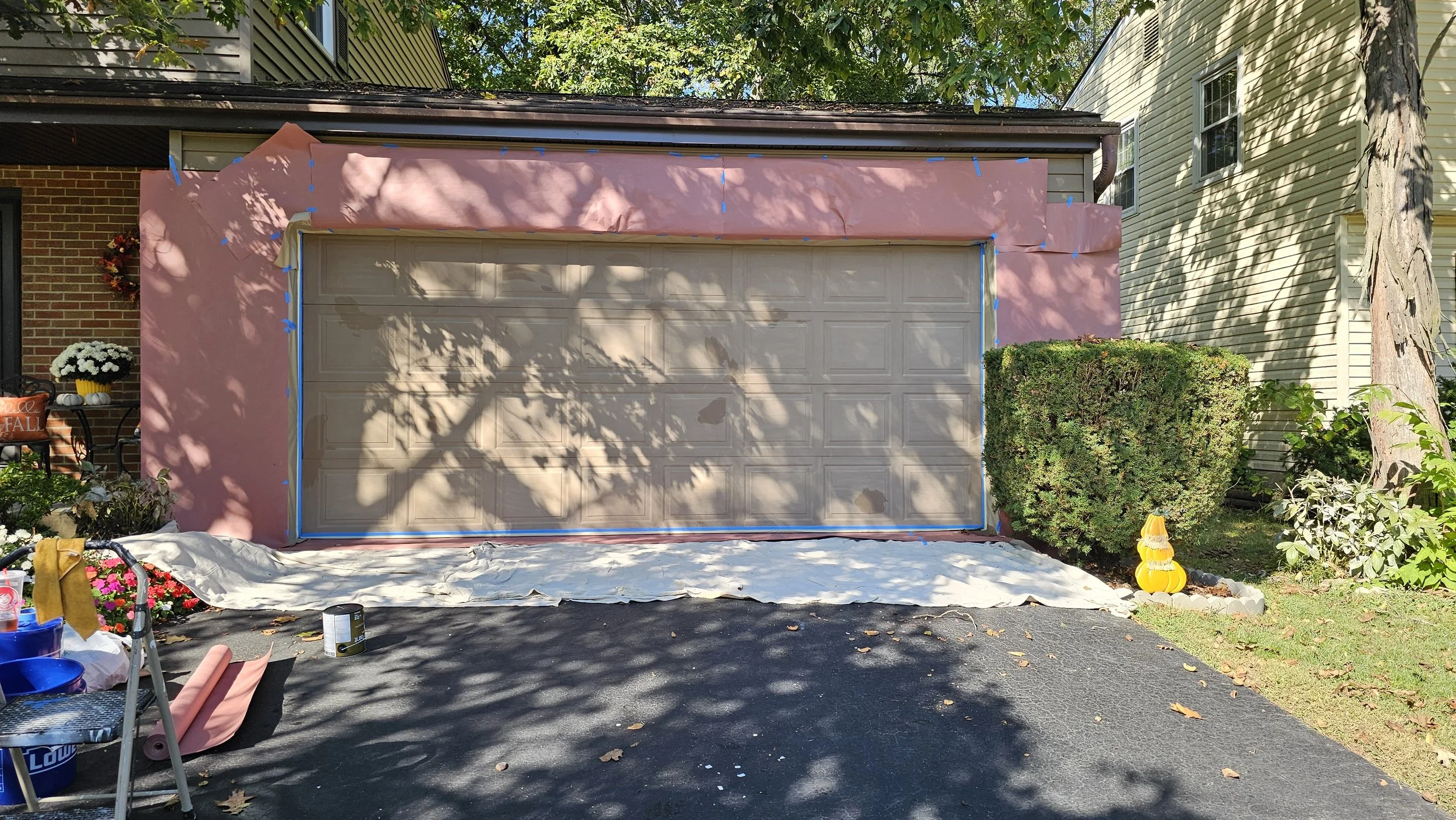 A house garage is being painted pink, with masking tape and tarps for painting protection. There are white flowers in a pot on a chair on the left, and a decorative yellow gourd sculpture on the ground near a bush on the right. The garage is surround