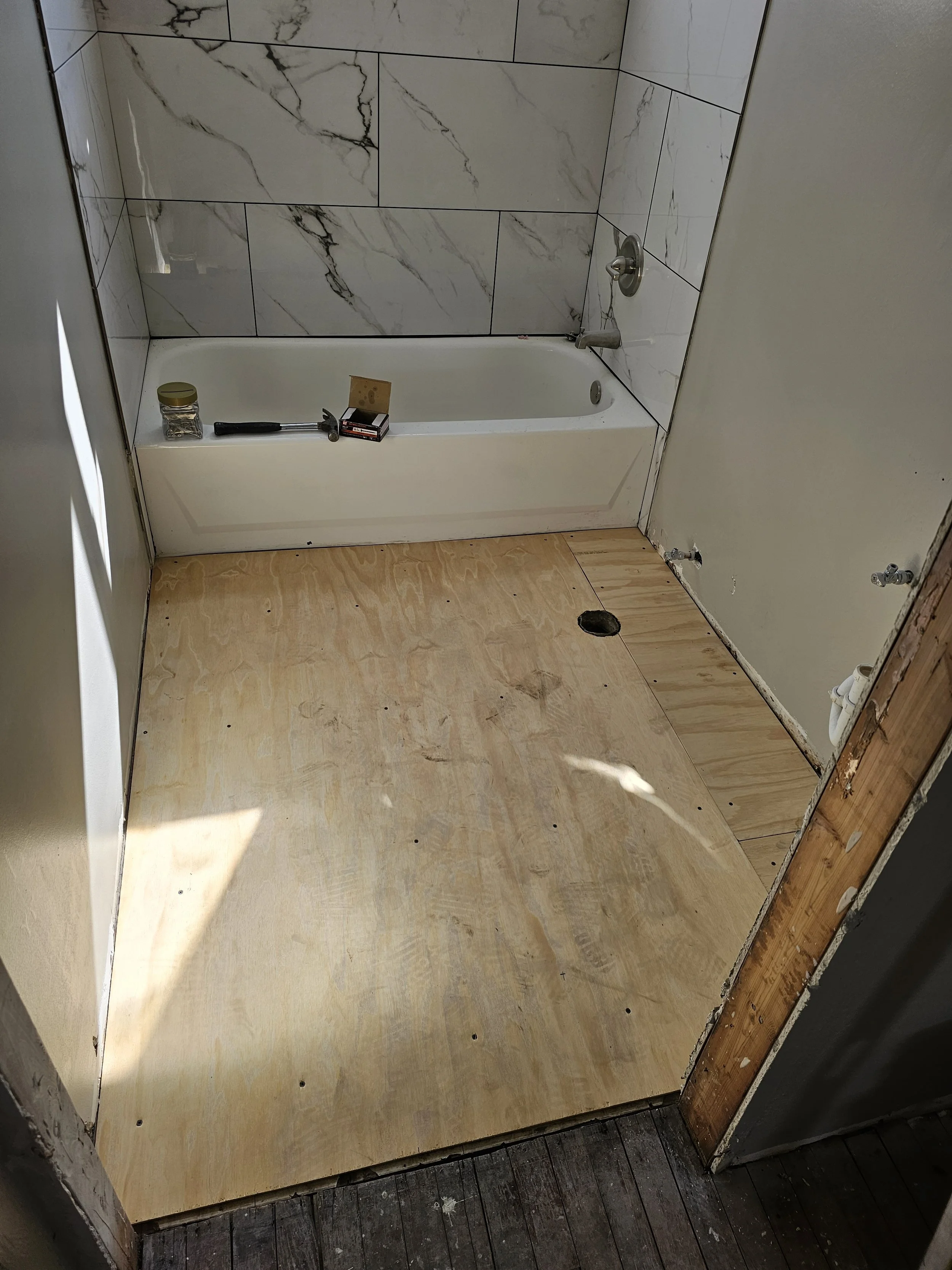 Bathroom under renovation with a bathtub, plumbing, and wood subfloor, with construction supplies on the edge of the tub.