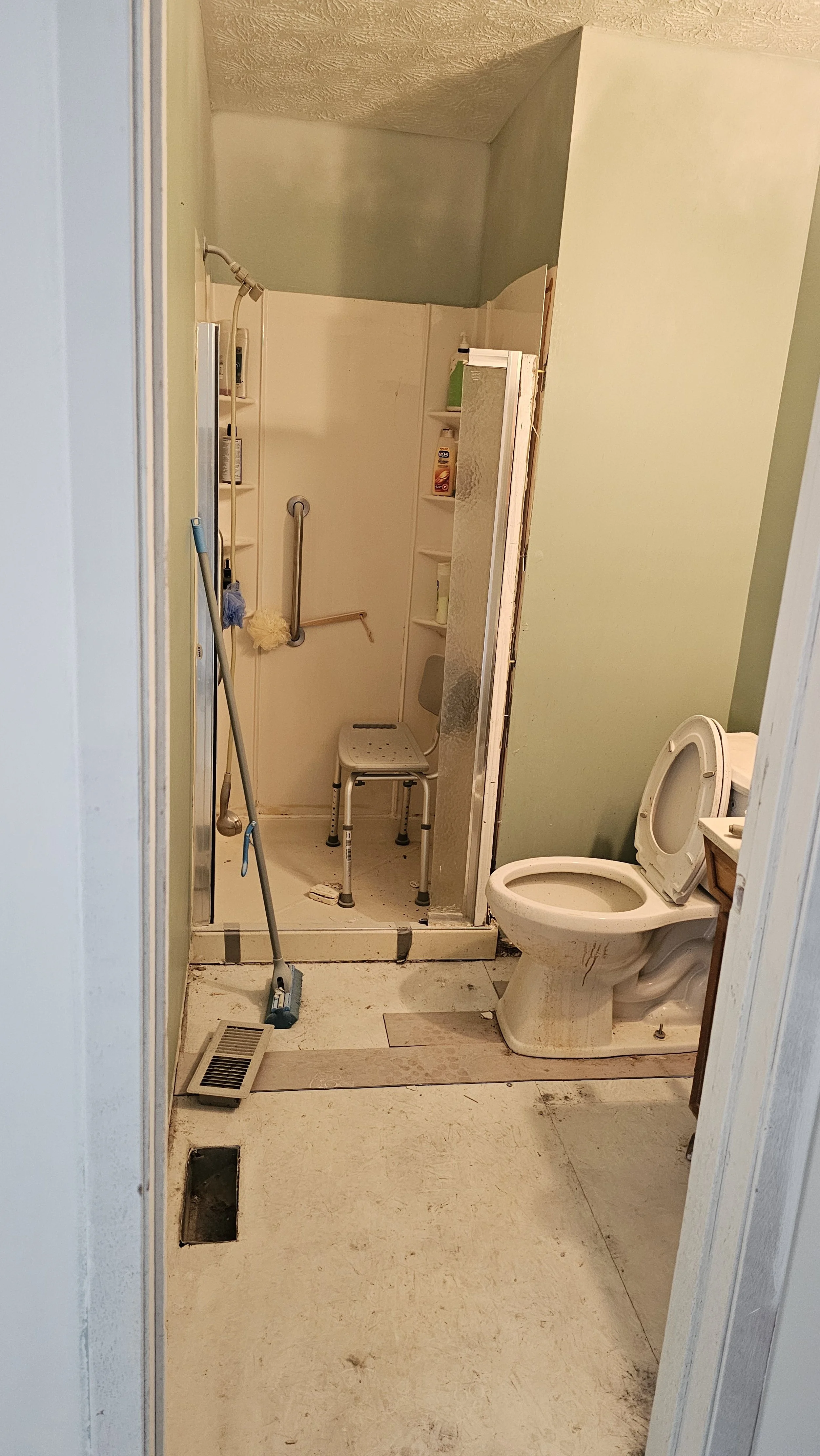 A small, cluttered bathroom with a toilet, shower area, and various cleaning supplies and tools. The floor is dirty and torn, and the shower is partially enclosed with a chair inside.