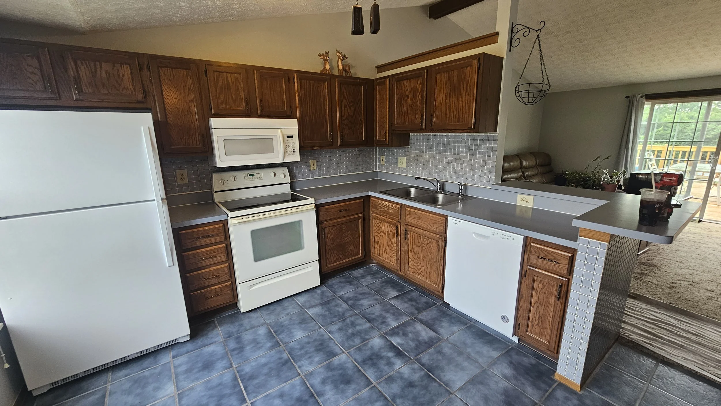 Kitchen with wooden cabinets, white appliances including refrigerator, microwave, and stove, gray countertop, small tiled backsplash, and a sink. The floor is tiled with dark blue tiles.