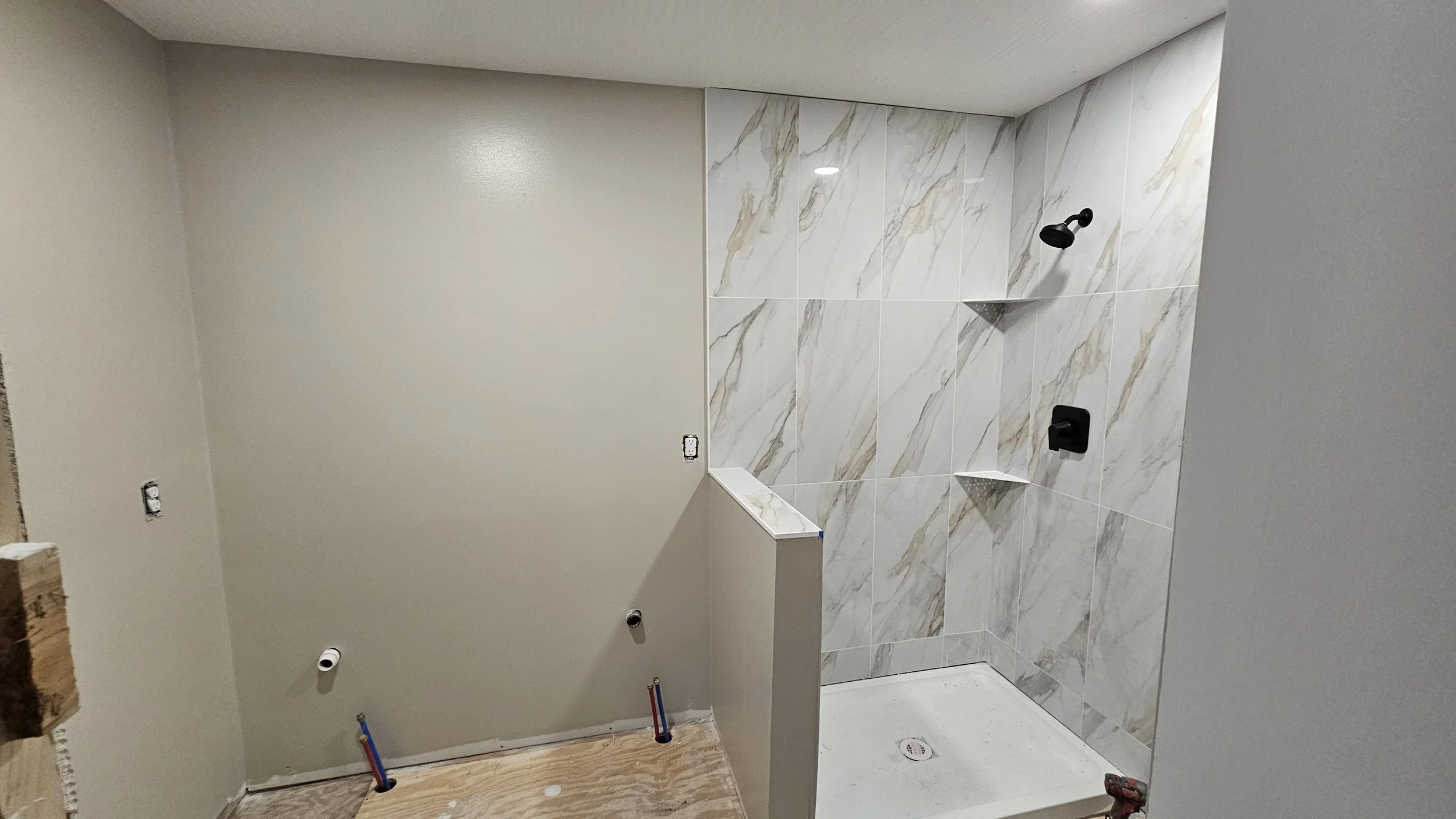 Bathroom shower area under construction with partially tiled wall, plumbing pipes, and a drain, but no fixtures installed yet.