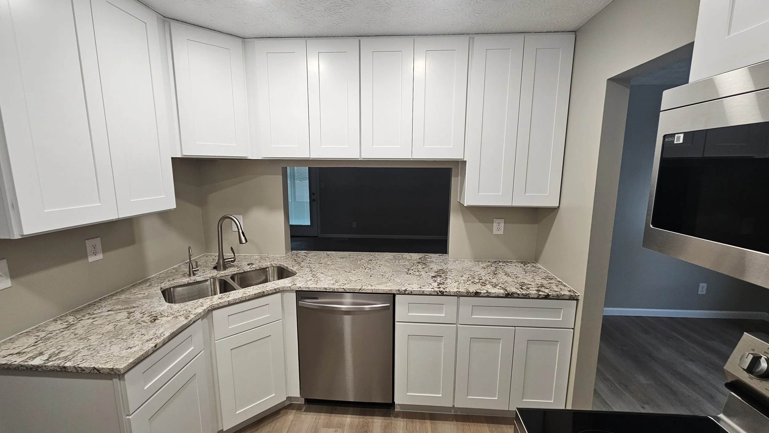 Kitchen with white cabinets, granite countertops, a stainless steel dishwasher, a double sink, and a pass-through window to the living area.