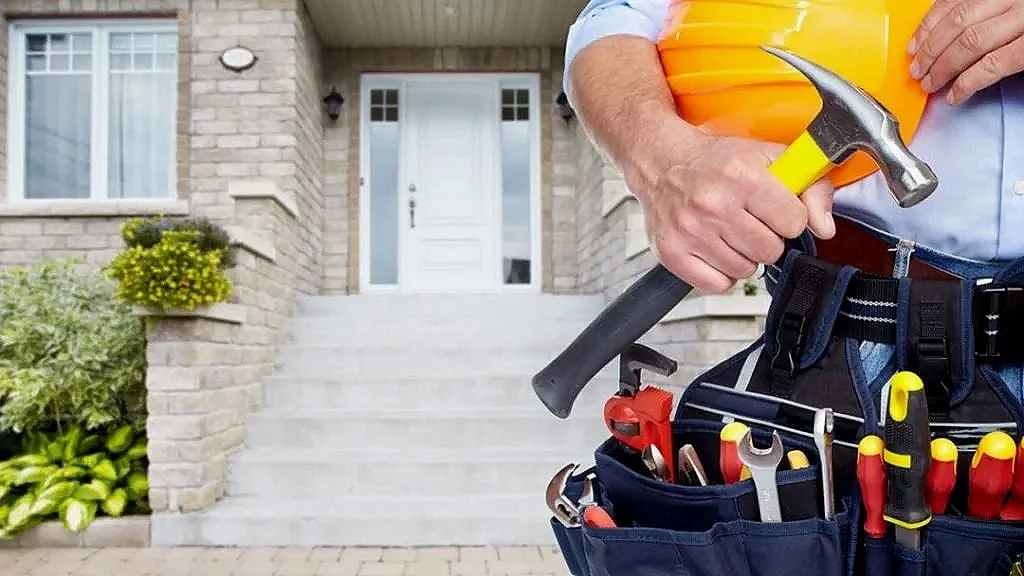 Close-up of a handyman holding a hammer and small pumpkin, wearing a tool belt with various tools, standing in front of a house staircase.