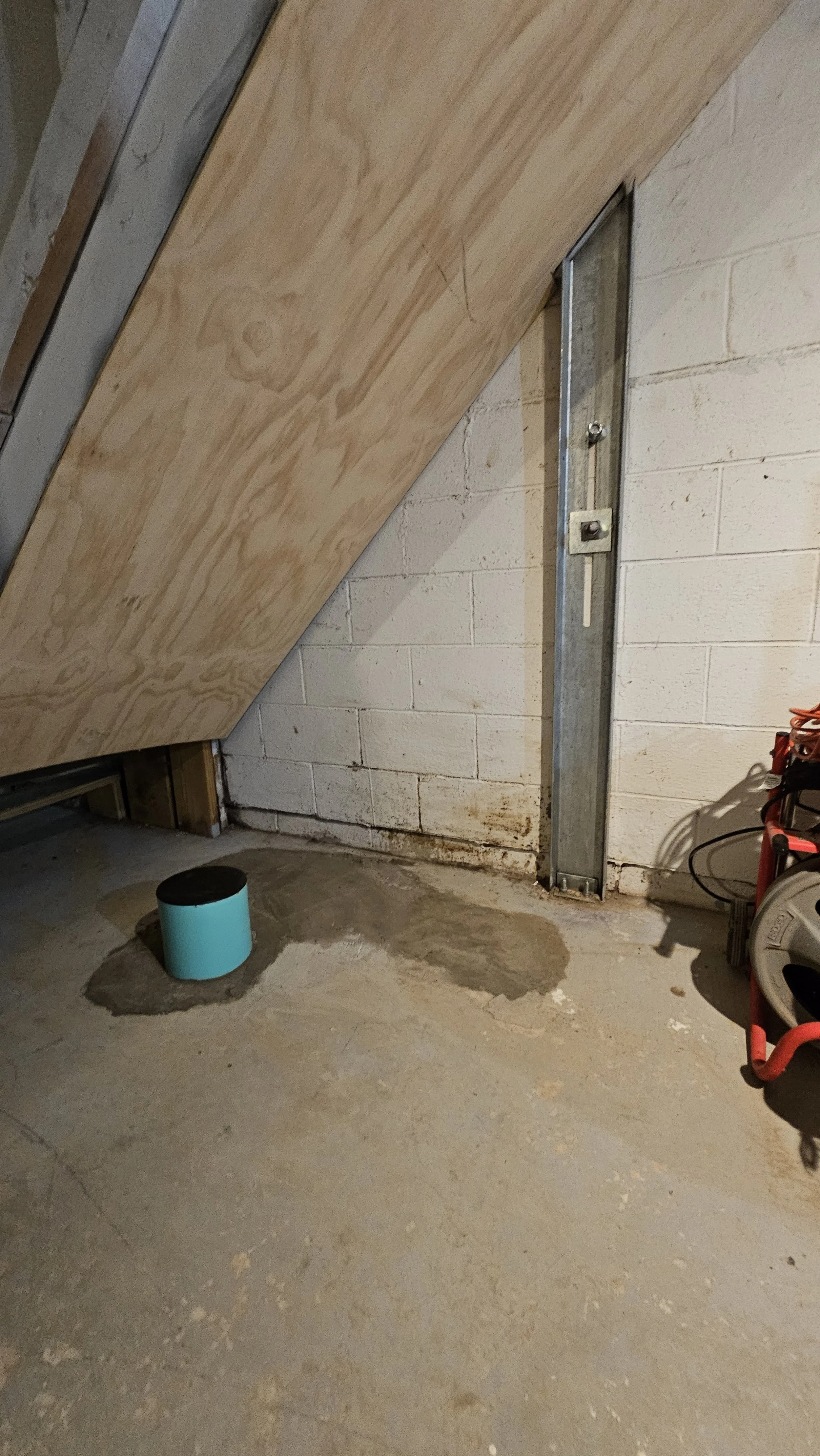 A garage with an unfinished interior, showing a sloped wooden ceiling, a cement floor with water stains, a metal support beam, and tools on the right side.