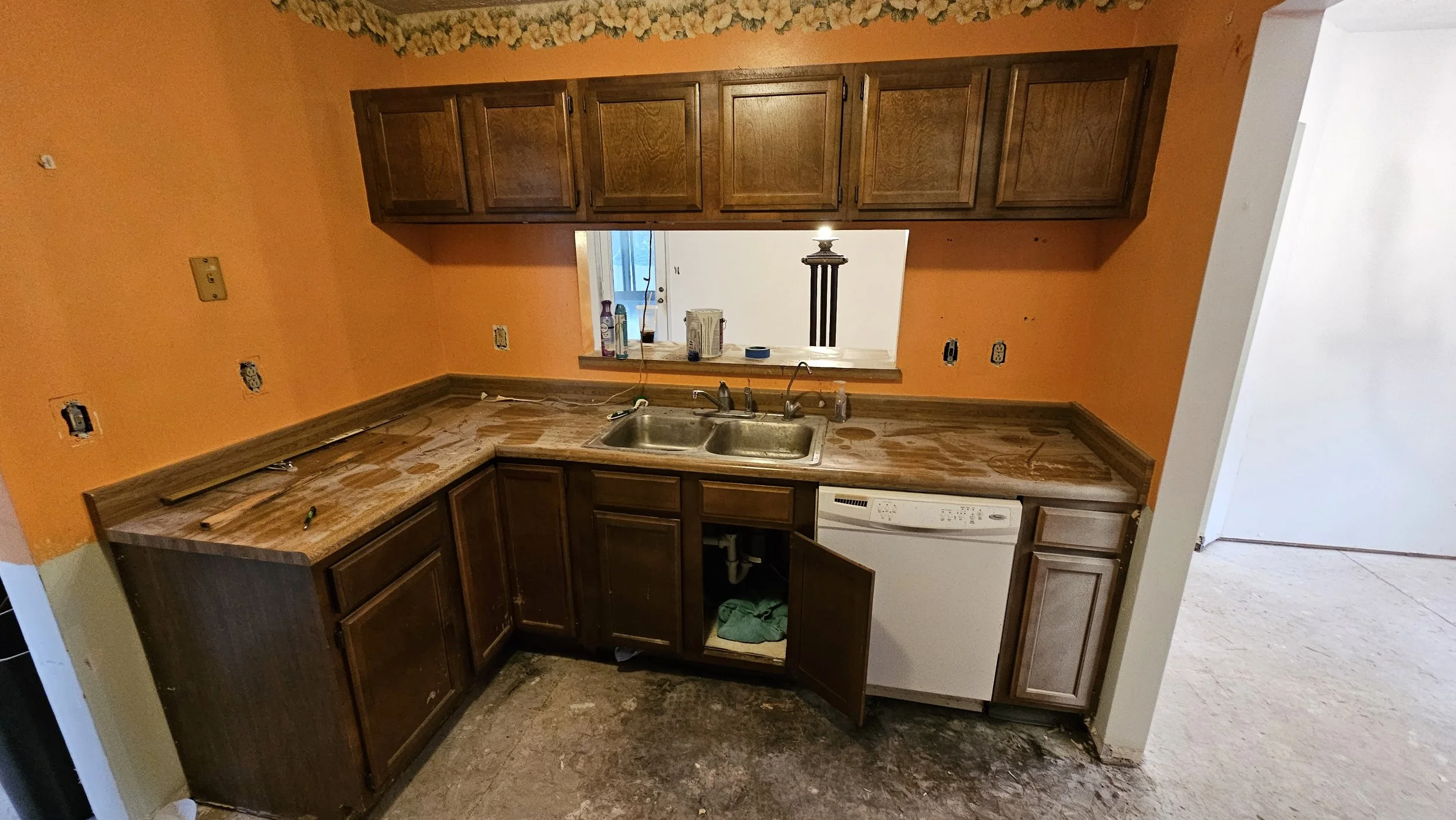 Kitchen with orange walls, dark wood cabinets, a double sink, and a dishwasher. The counter is cluttered with cleaning supplies, and the space appears to be under renovation, with missing wall outlets and a messy floor.