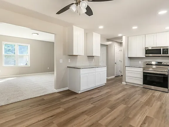 Kitchen with white cabinetry, stainless steel stove and microwave, wood flooring, ceiling fan, and open to a living area with carpet and large windows.
