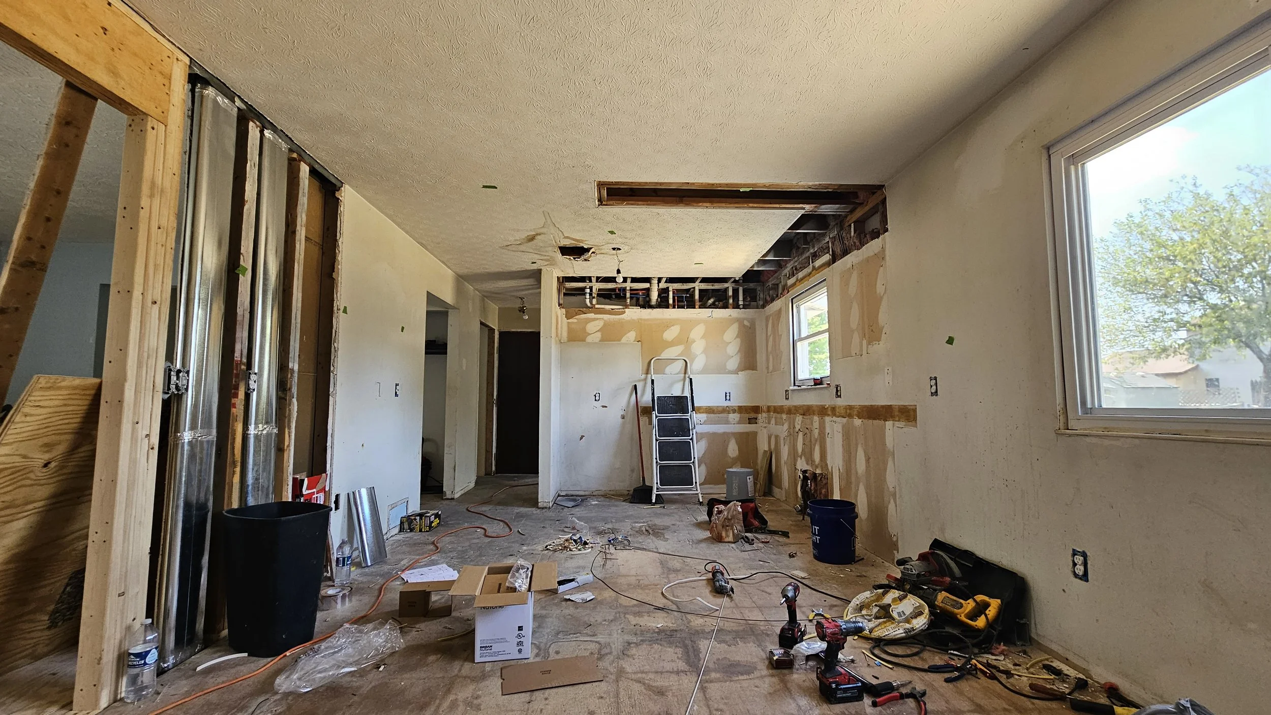 Interior of a house under renovation with exposed walls and ceiling, construction tools scattered on the floor, a ladder, and a window showing trees outside.