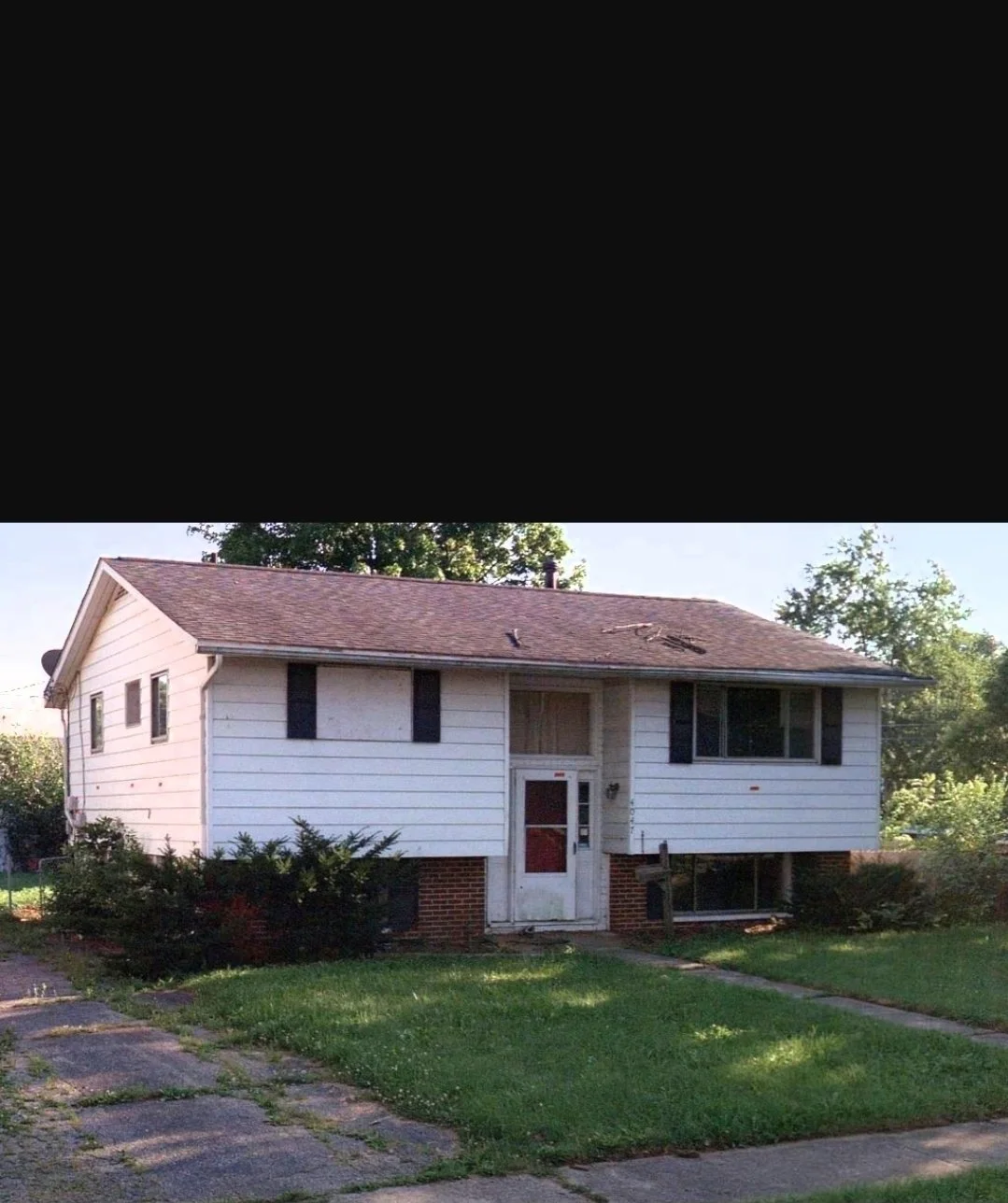 A two-story house with white siding, black shutters, and a brick foundation. The front door is boarded up, and the windows are covered or broken. The yard is grassy with some shrubs, and there is a walkway leading to the front door.