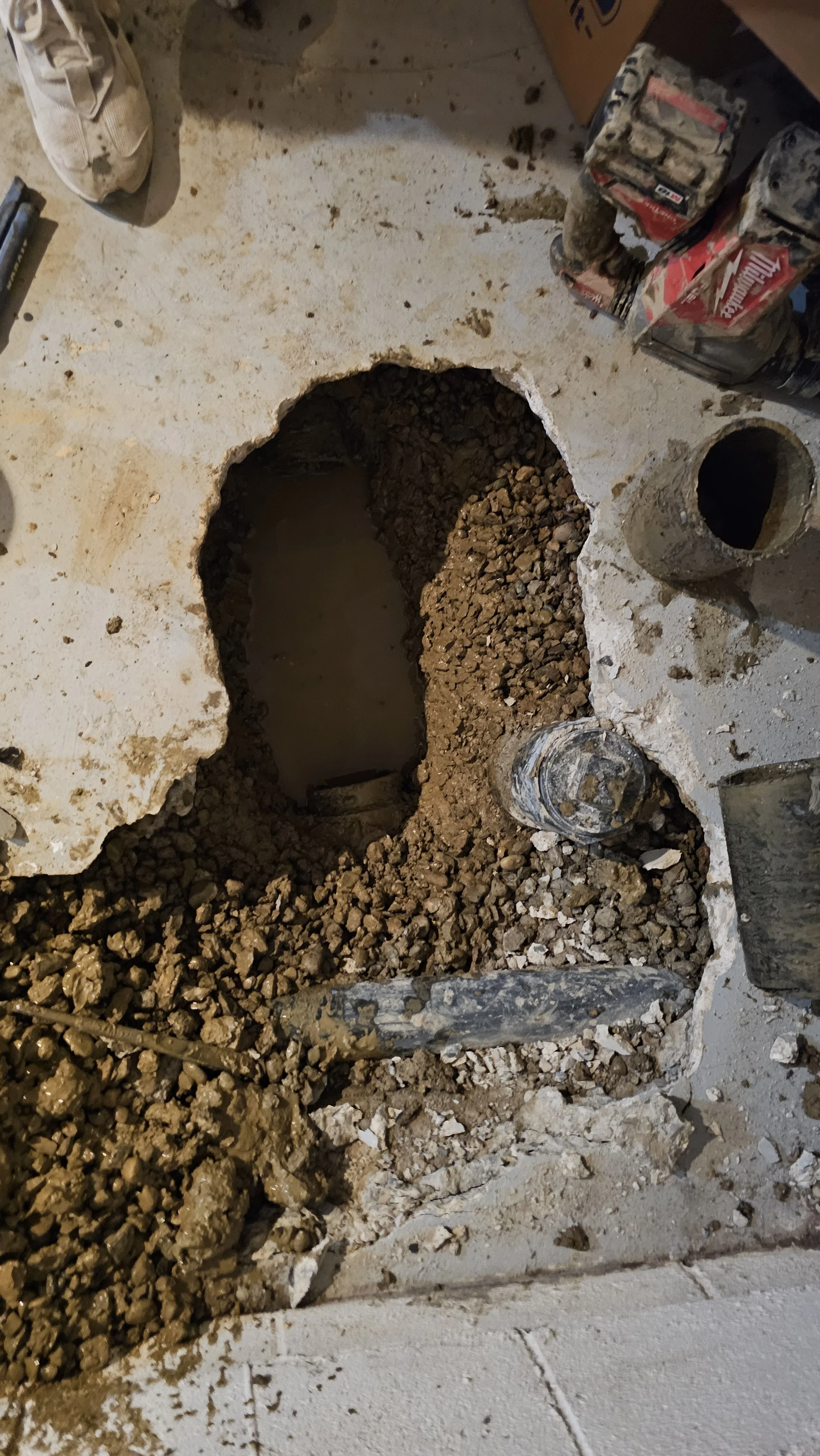 An open hole in the concrete floor reveals a pipe and some gravel inside, surrounded by construction tools and materials.