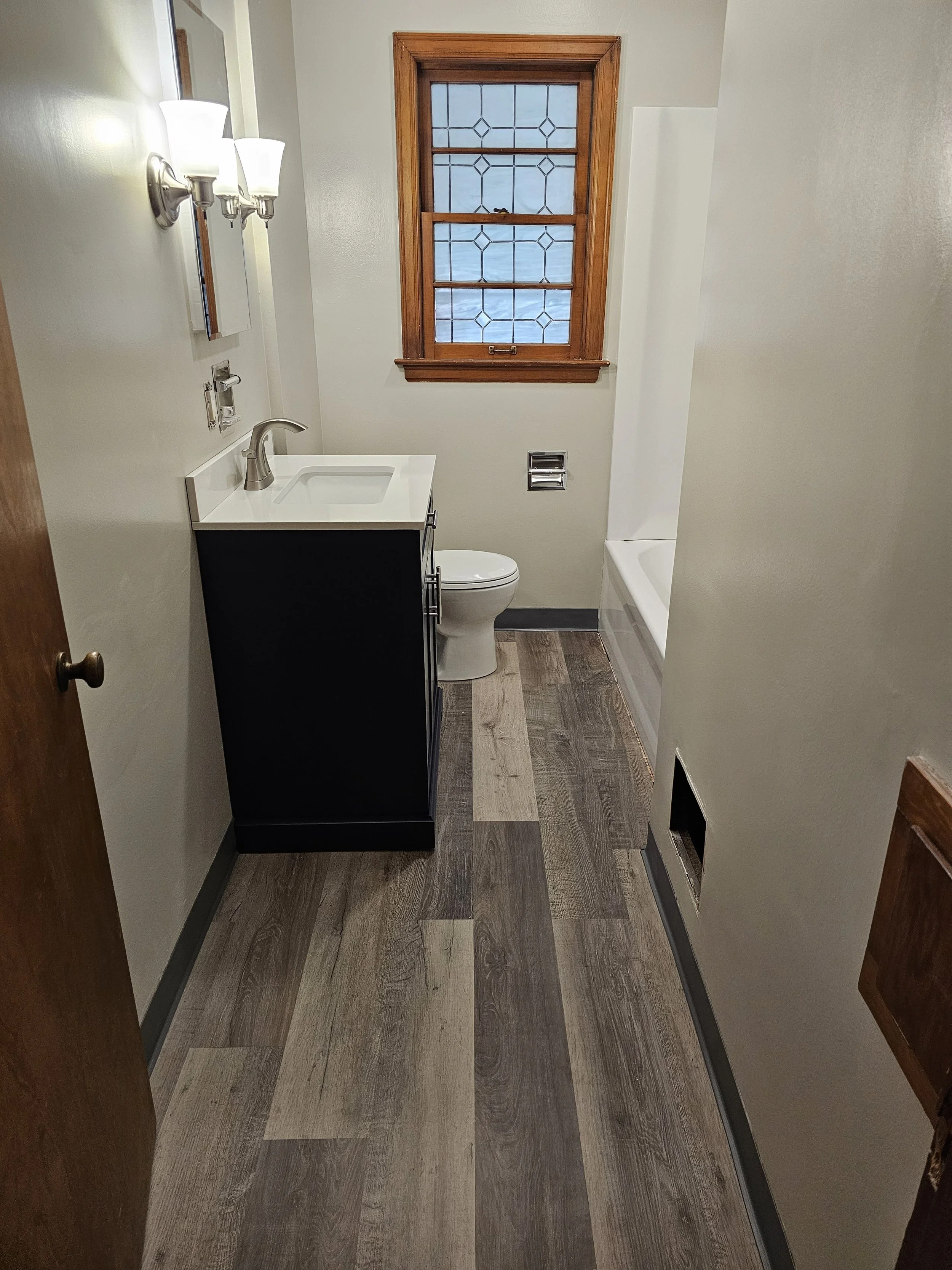 Small bathroom with a vanity with a white sink, toilet, bathtub, wood-framed window, and wood flooring.