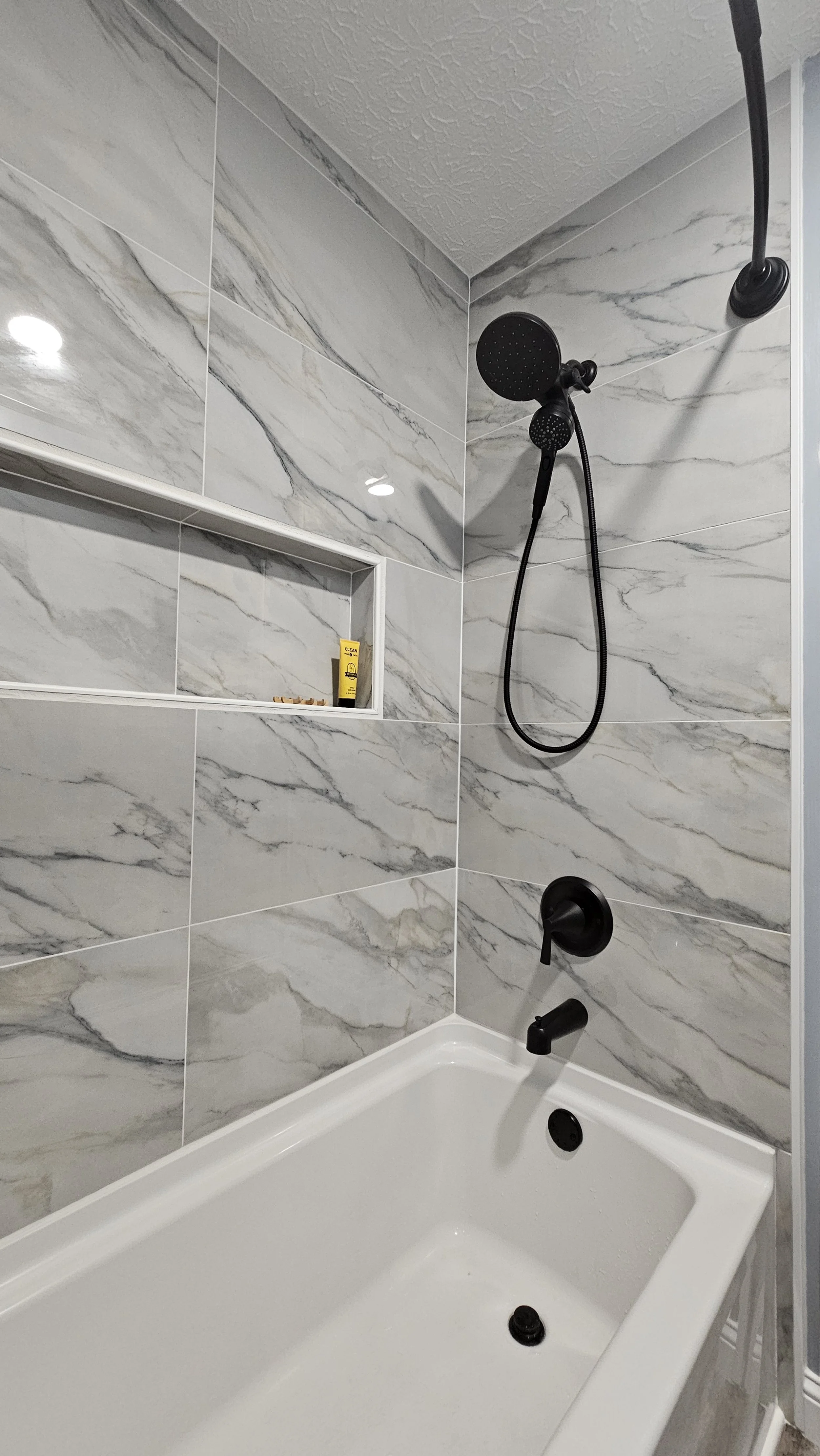 Bathroom shower with black fixtures, marble tiles, niche with toiletries, white bathtub, and shower head.