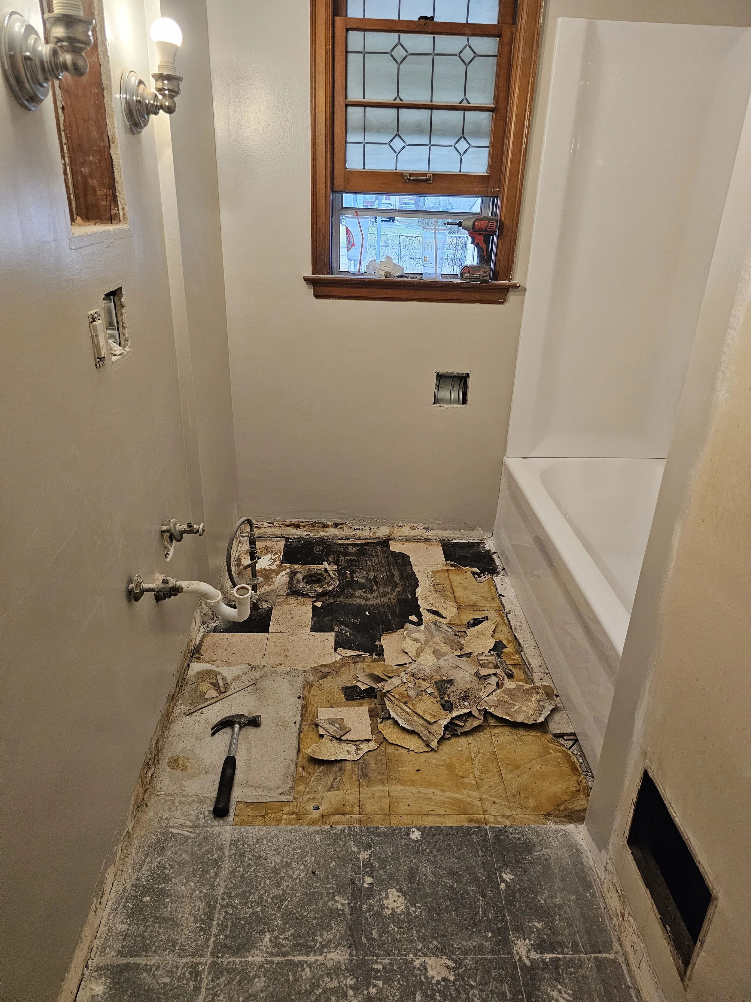 A bathroom under renovation with exposed plumbing, damaged flooring, and removed tiles. There is a window with a cordless drill on the sill, and debris on the floor, including torn paper and broken tiles.
