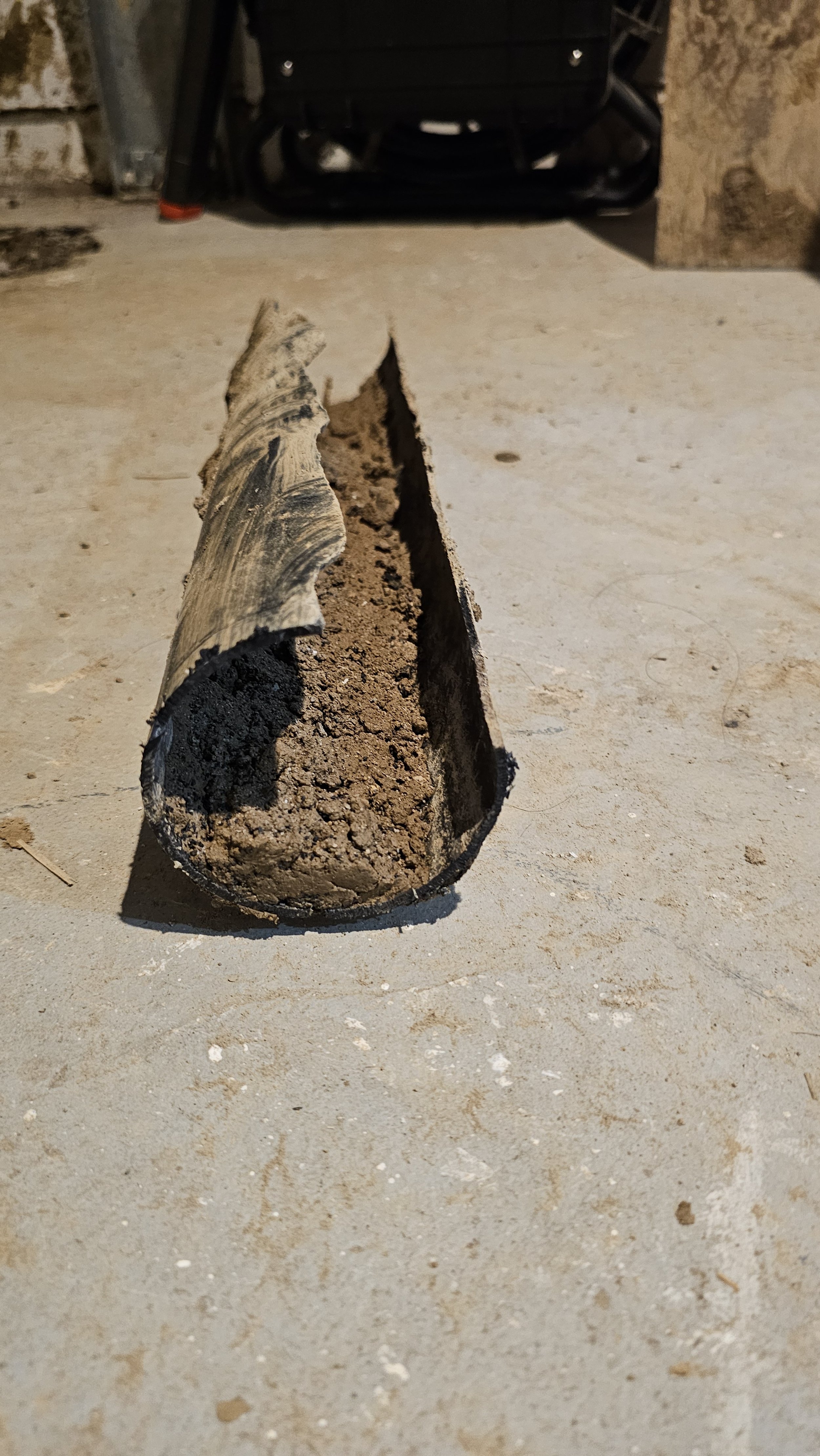 Close-up of a cut section of a metal pipe filled with dirt, placed on a concrete floor in a garage or basement.