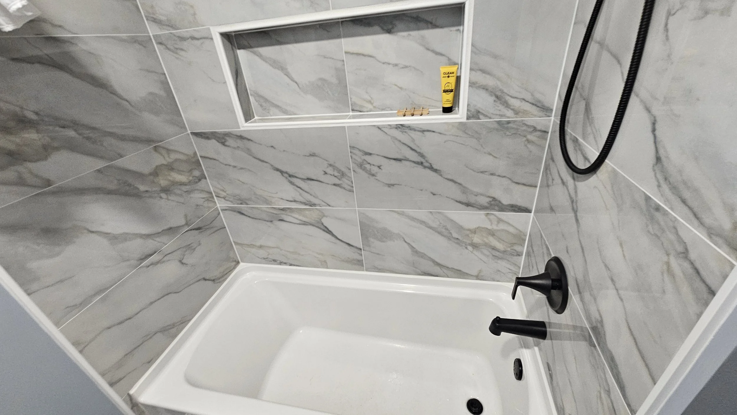 Empty bathroom shower with marble tiled walls, a built-in niche with a tube of shampoo, a small decorative item, black shower fixtures, and a black shower hose.