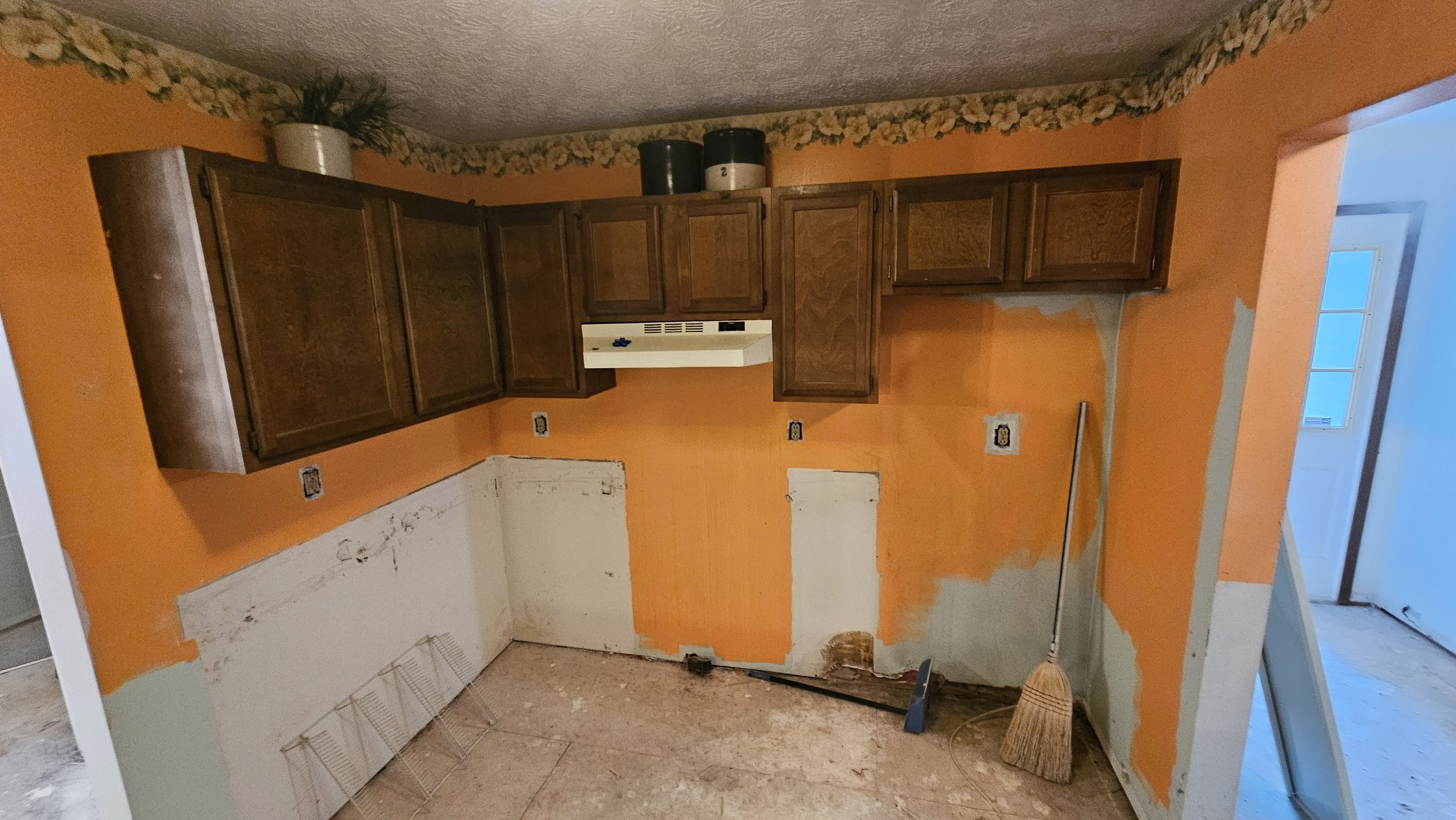 Kitchen with orange walls and wooden cabinets, partially painted walls with visible primer and patchwork, broom and dustpan on the floor, and an open door leading to another room.