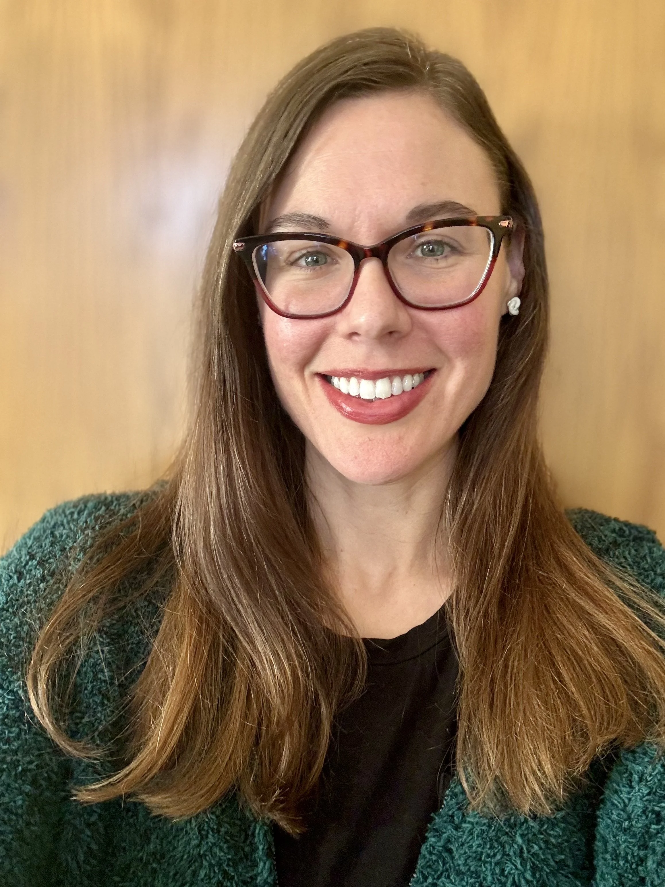 A woman with long brown hair, wearing glasses, earrings, a black top, and a green textured jacket, smiling against a brown background.