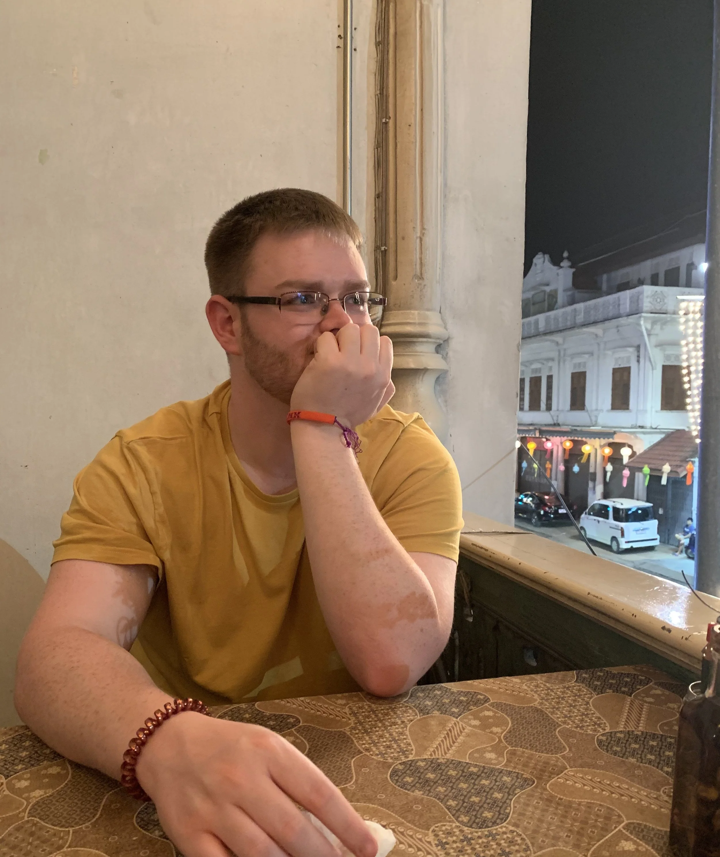 A young man with glasses sitting at a table, resting his chin on his hand, looking out the window at night, with colorful lanterns and buildings visible outside.