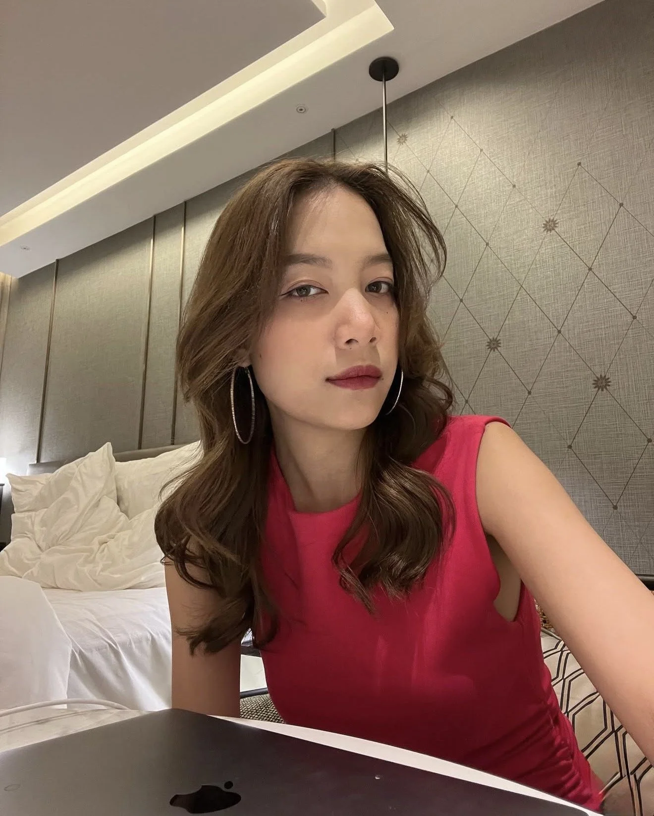 A woman with wavy brown hair wearing large hoop earrings and a pink sleeveless top, sitting on a bed in a modern bedroom with beige and gray decor, and a laptop in the foreground.