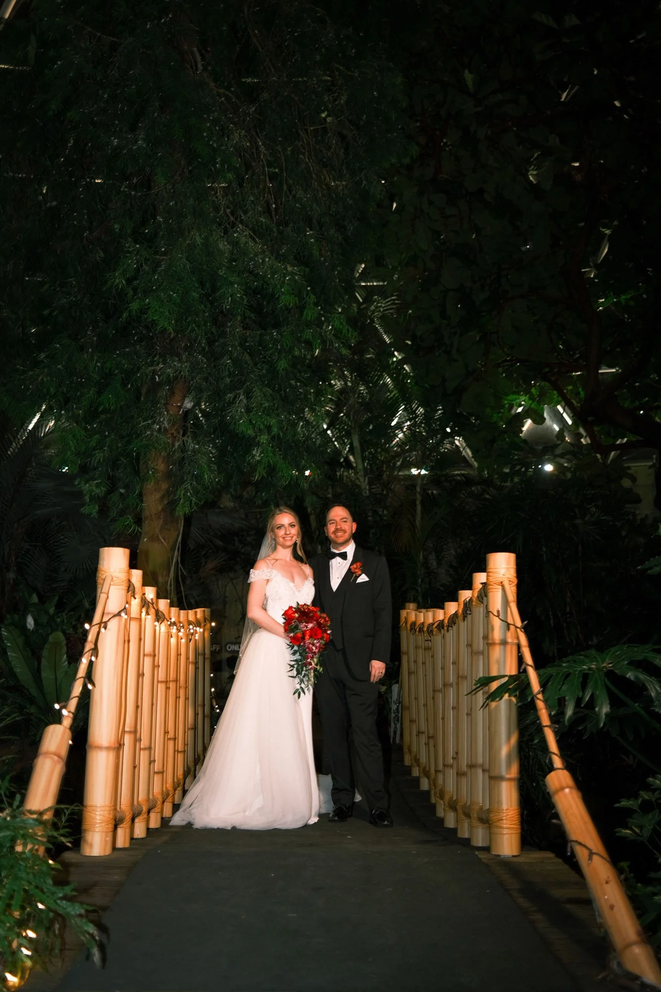 A bride and groom standing together on a wooden bridge at night, surrounded by lush greenery. The bride is wearing a white wedding gown and holding a bouquet of red flowers. The groom is dressed in a black tuxedo with a bow tie and orange boutonniere. They are smiling.