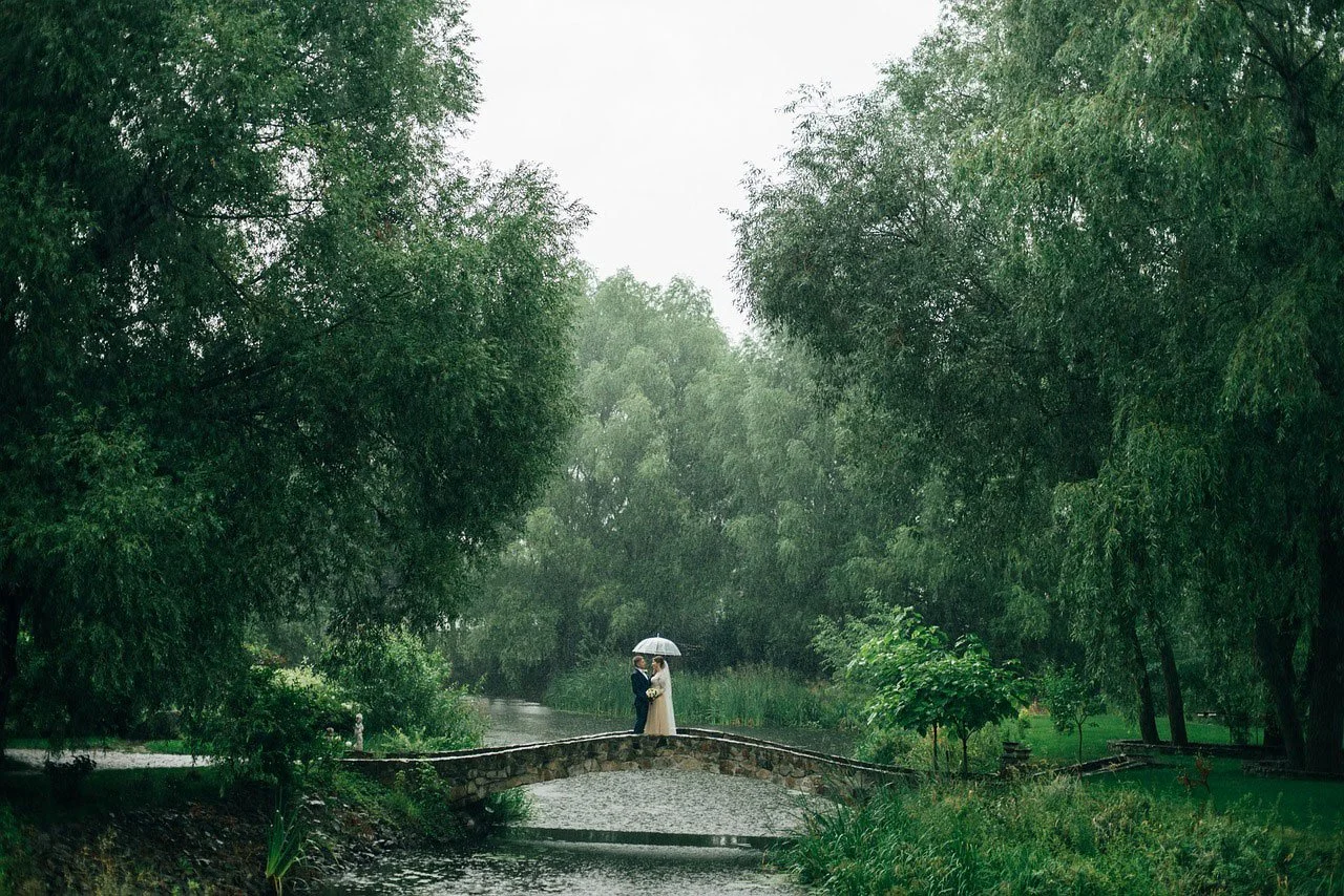 A bride and groom standing on a small stone bridge over a river in a lush, green park during rainy weather, with the bride holding a white umbrella.