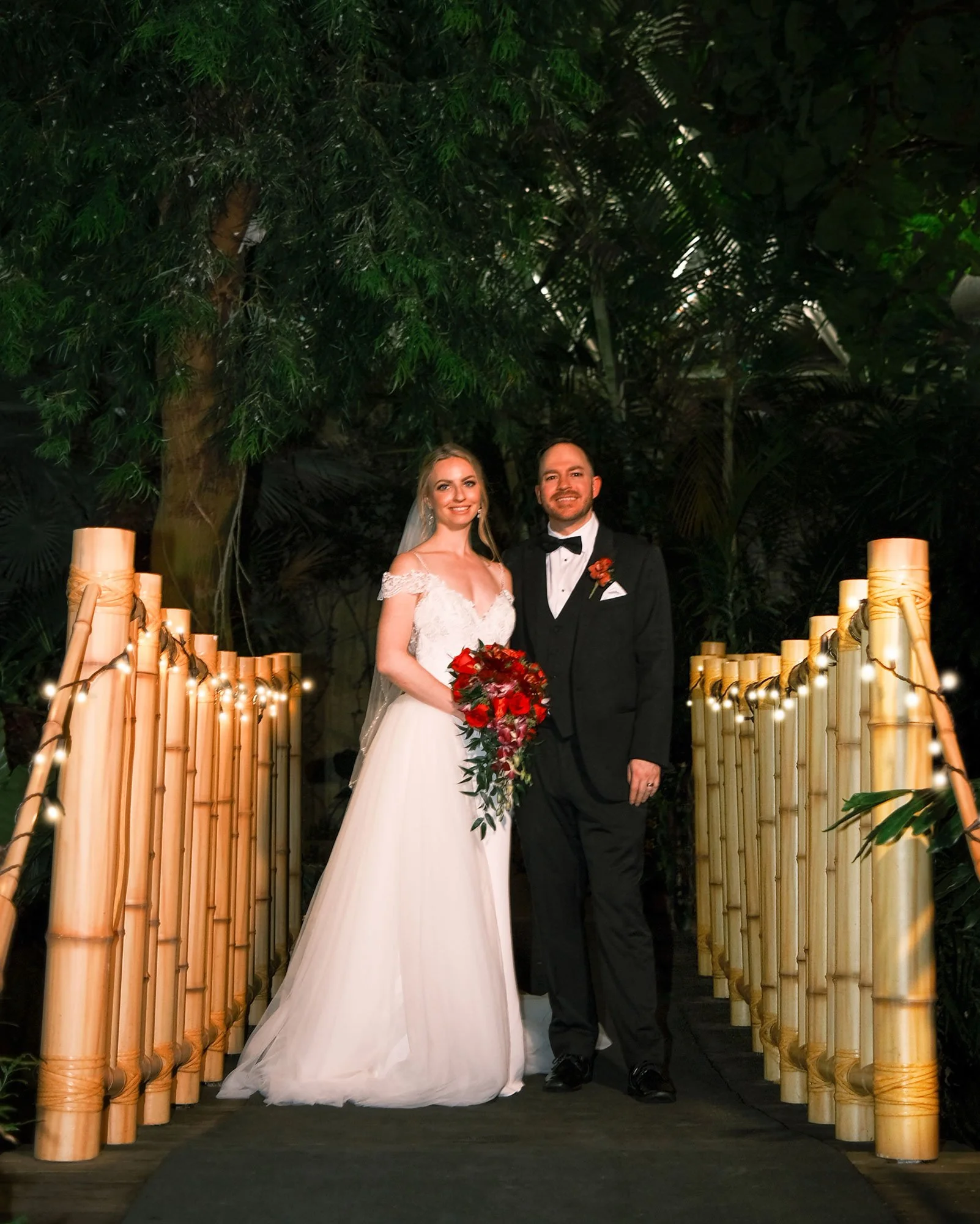 A bride and groom standing on a walkway decorated with bamboo and string lights, posing for a wedding photo in a lush, green outdoor setting at night.