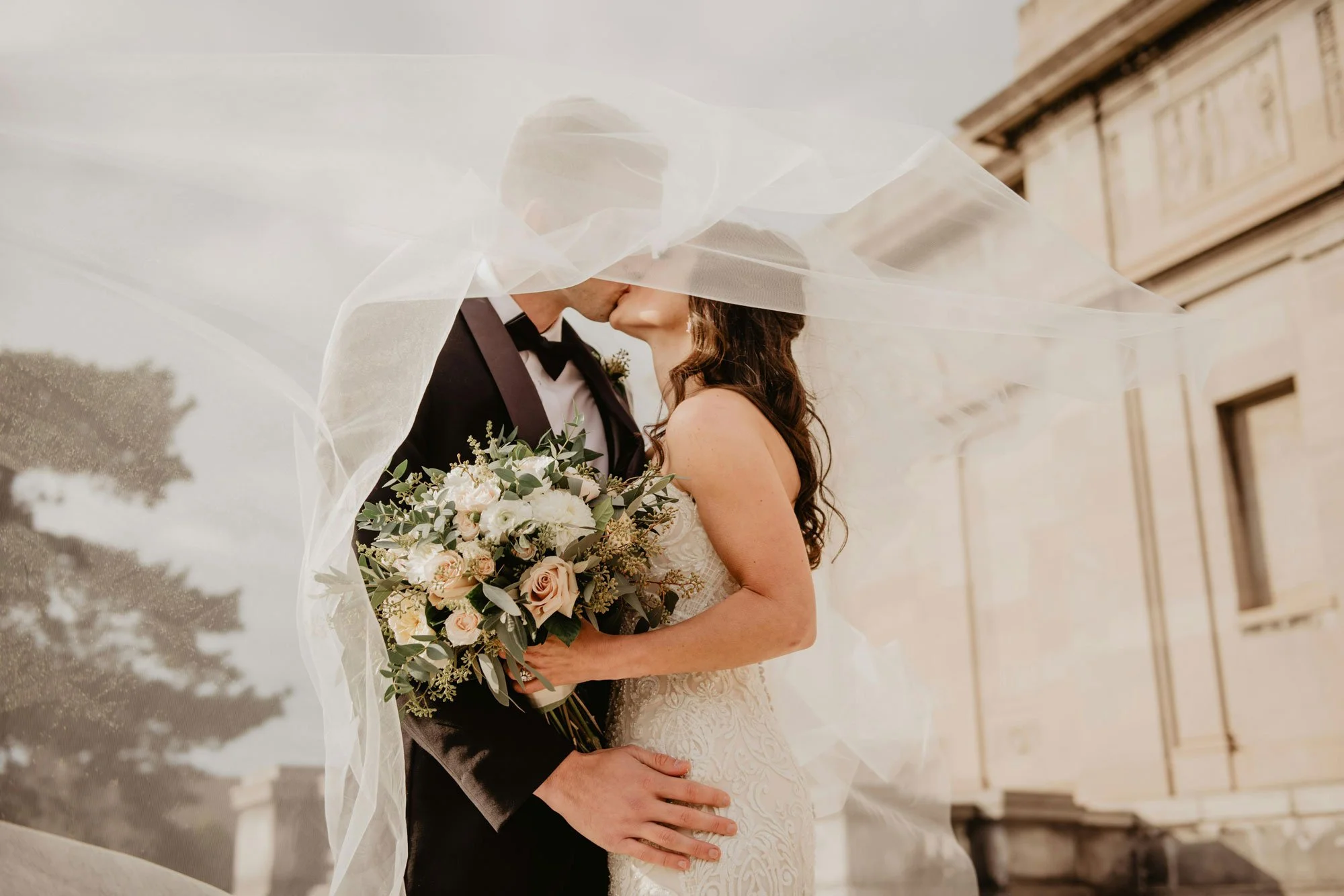 A bride and groom share a kiss under a flowing veil on their wedding day. The bride holds a bouquet of white and blush flowers. They stand outside a historic building.