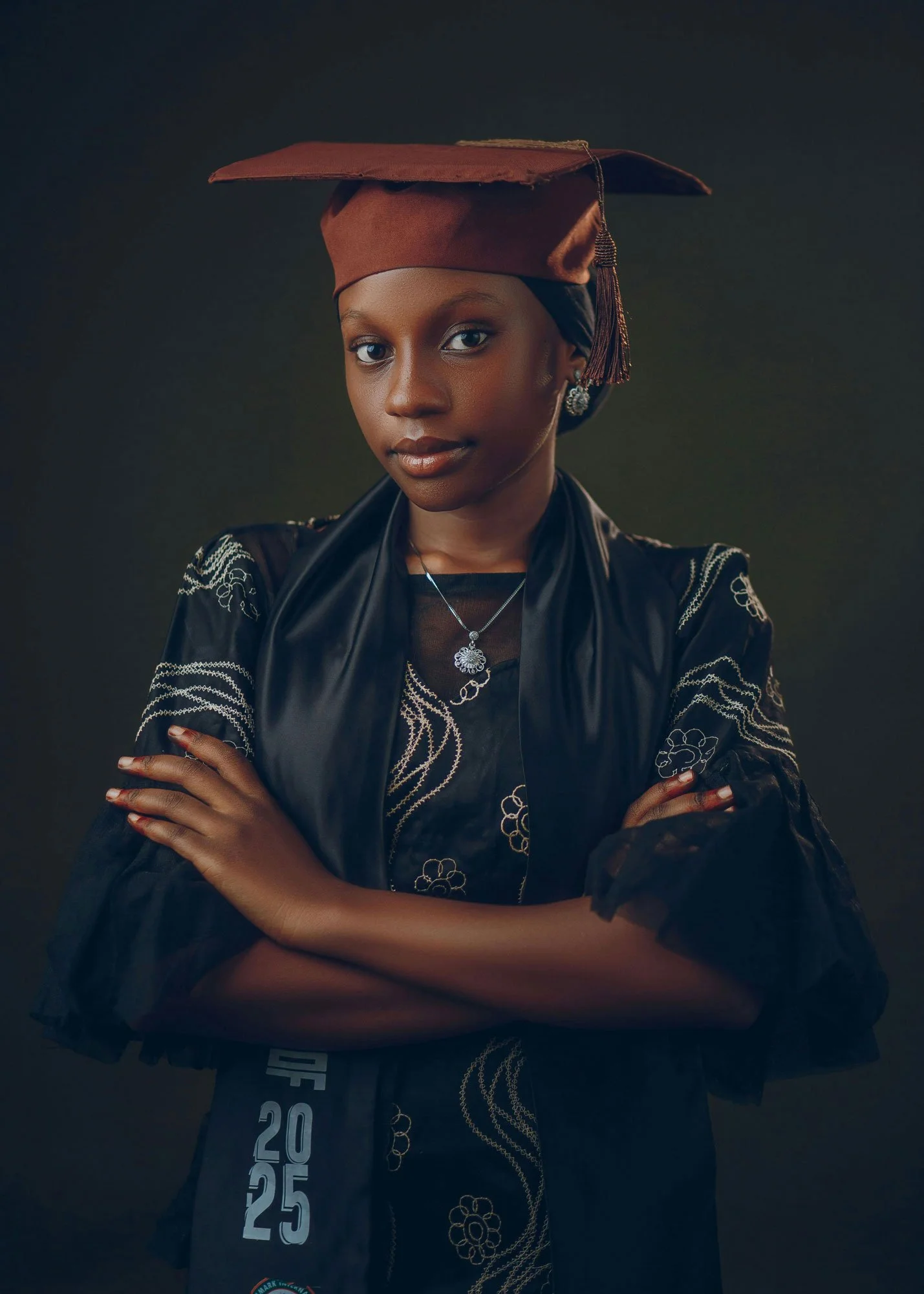 Studio graduation portrait of a young woman wearing a cap and gown with arms crossed against a dark background.