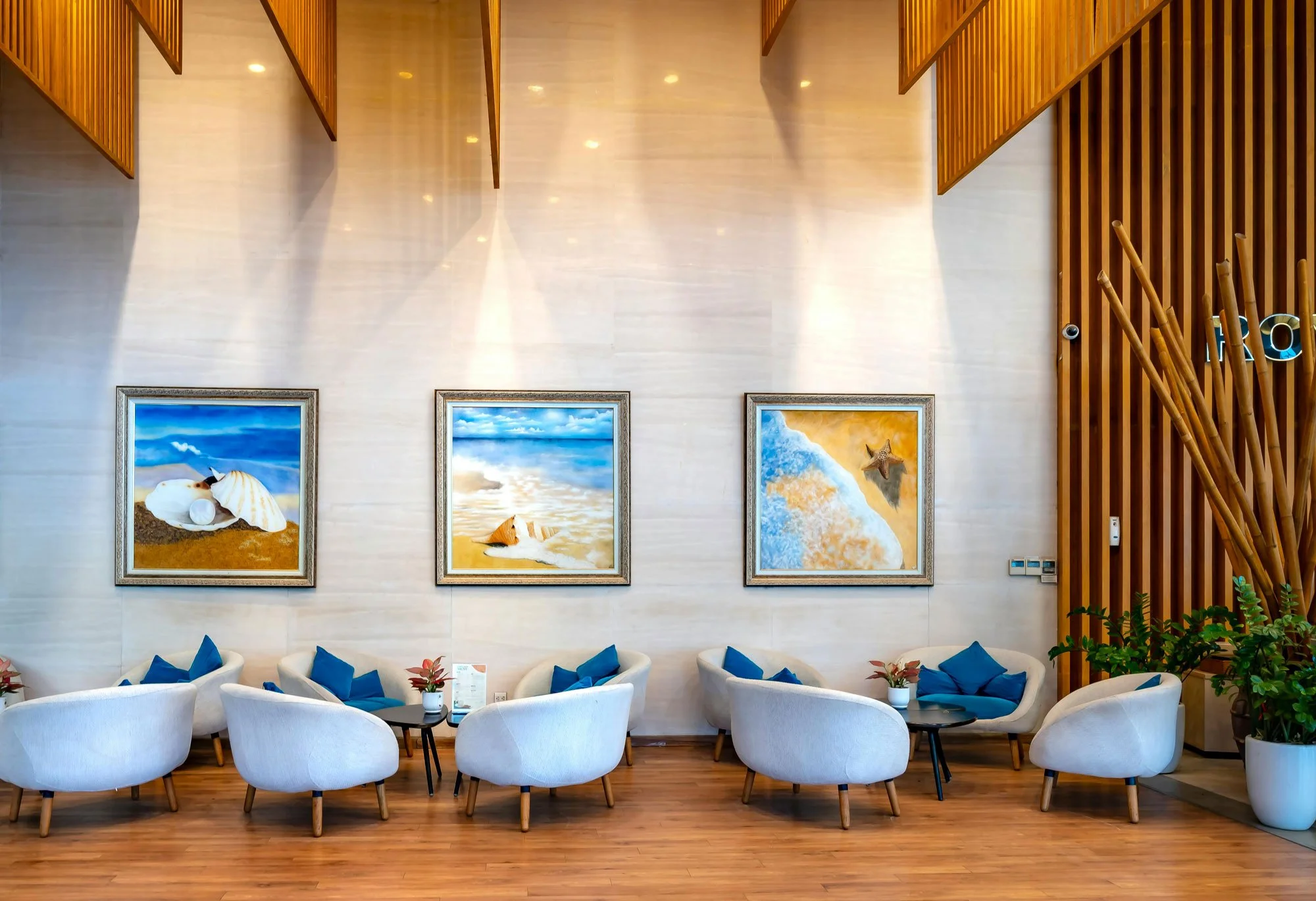Beach-themed paintings hanging on a cream-colored wall in a hotel lobby with white curved armchairs, small black tables, and potted plants.