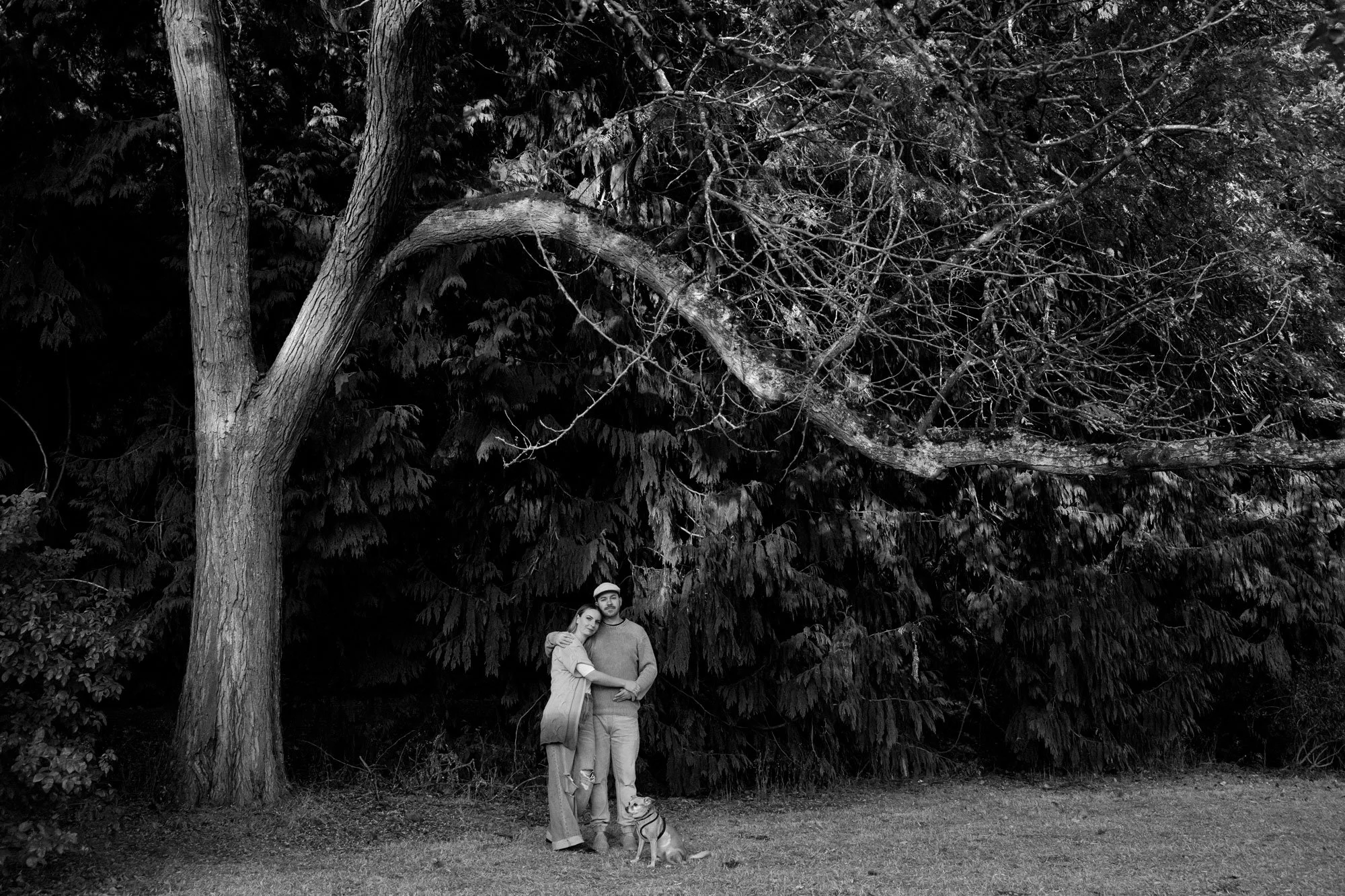 A couple standing with their dog in a park with tall trees and dense foliage in the background, in black and white.