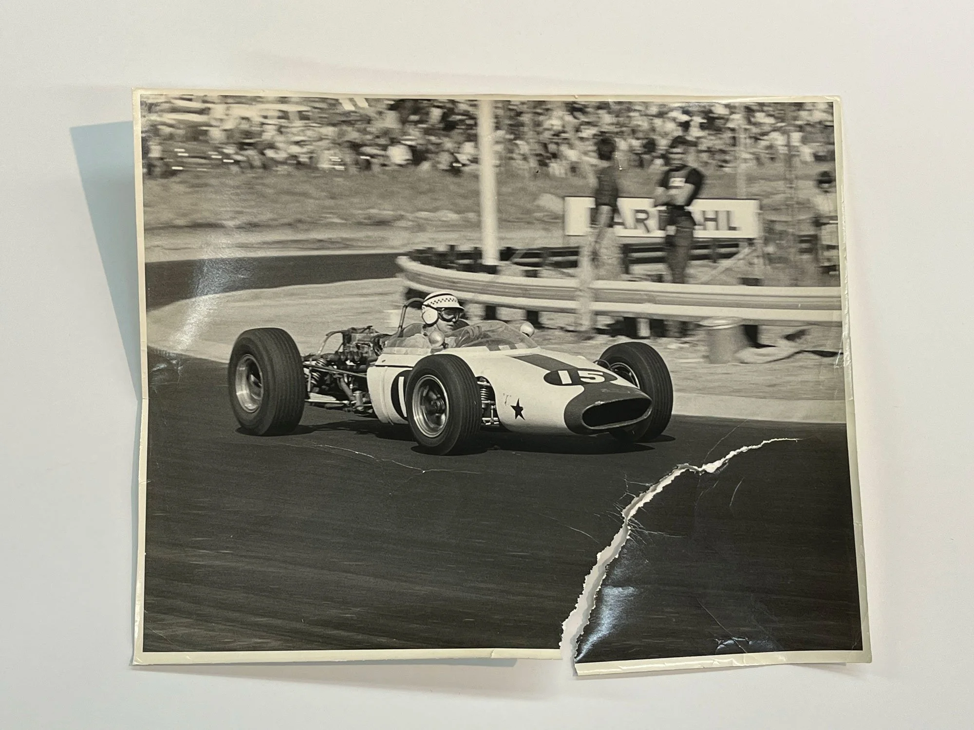 A black and white photograph of a vintage race car on a racetrack with a driver wearing a helmet and goggles, a crowd of spectators in the background, and some standing officials or crew members nearby.